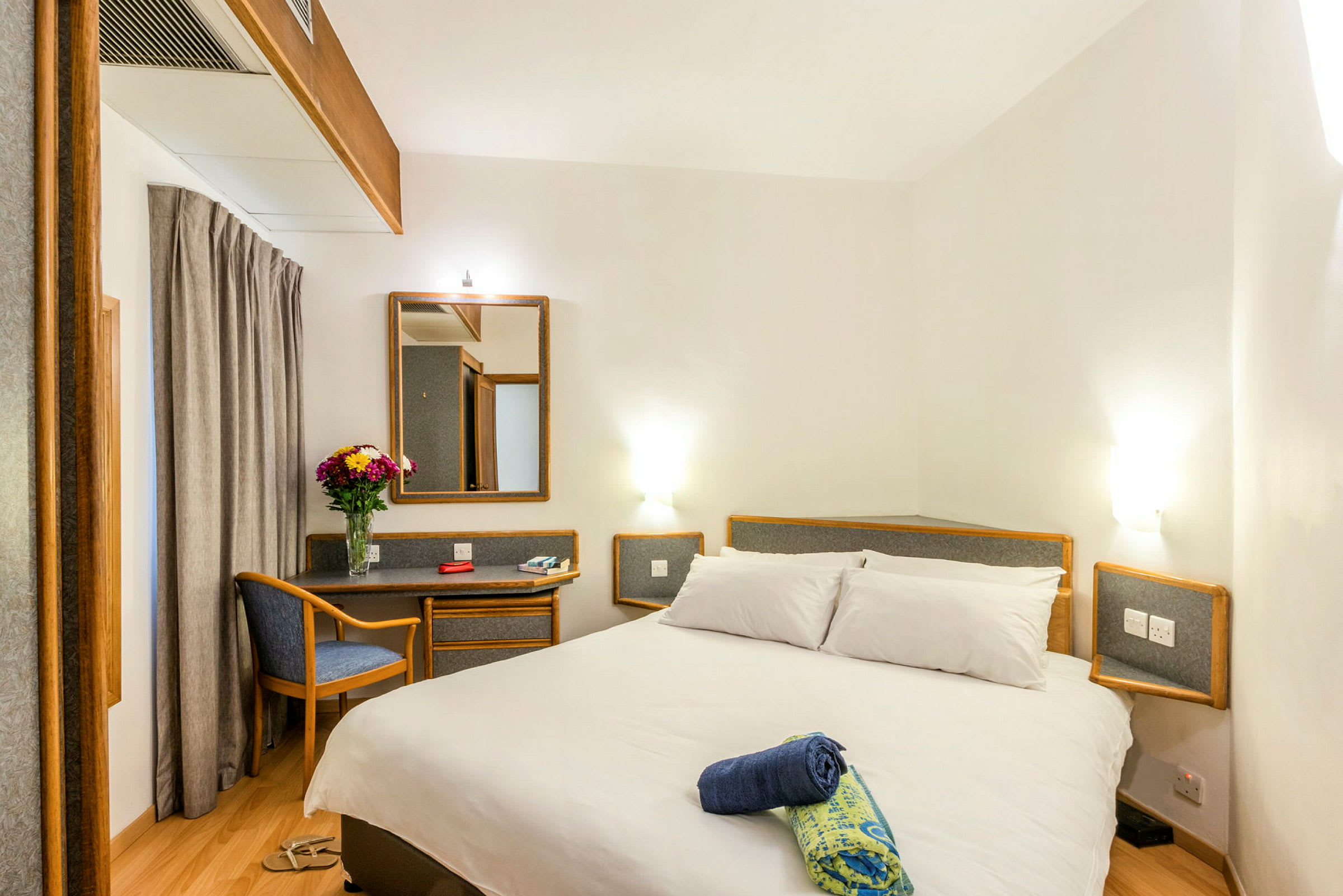 Ett hotellrum med en dubbelsäng, en spegel, en liten skrivbord och en stol. På skrivbordet står en vas med blommor. (AI-genererad text)