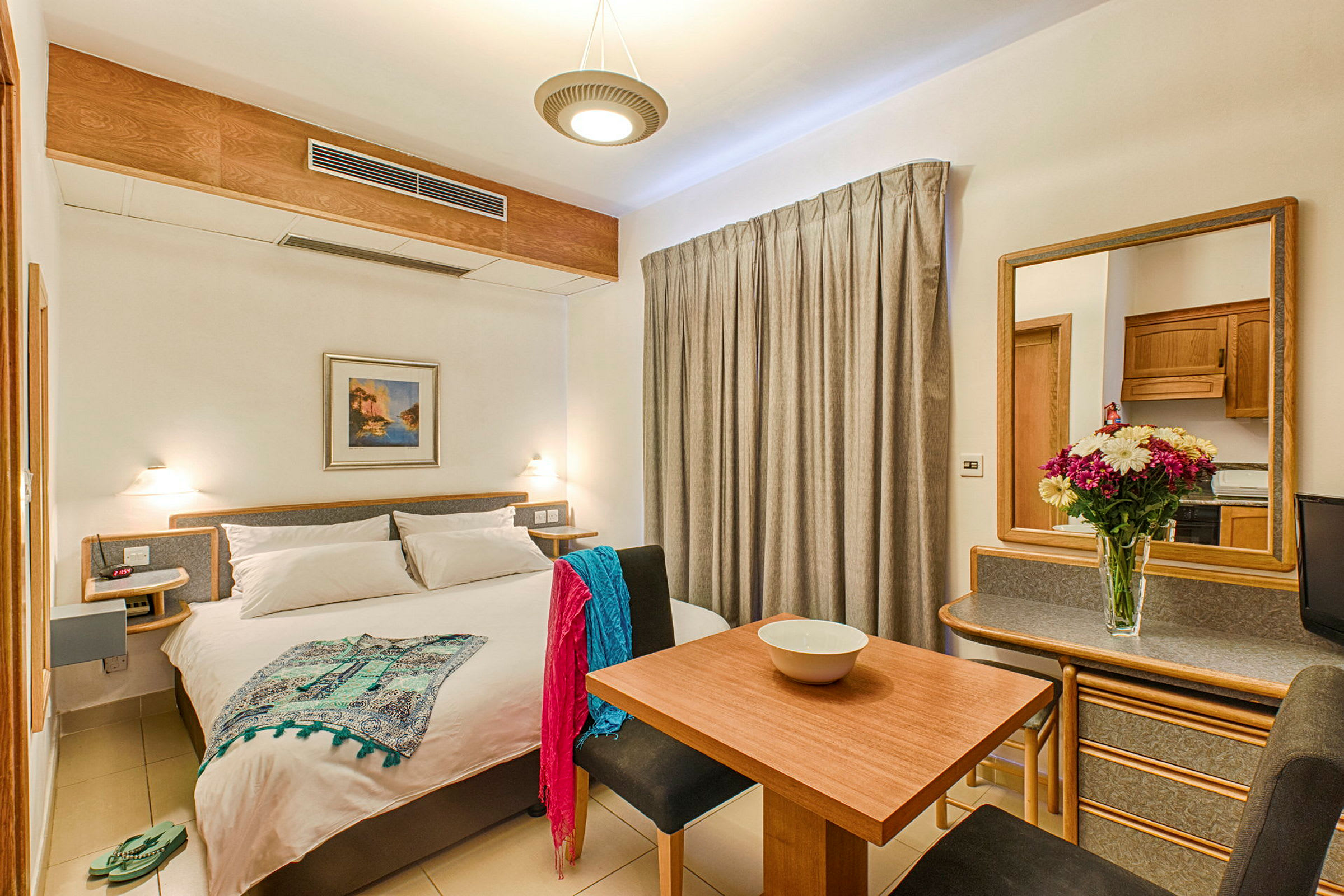 Ett ljust hotellrum med en dubbelsäng, matbord, sittplatser och en spegel. Dekorerat med en blomvas på bordet. (AI-genererad text)