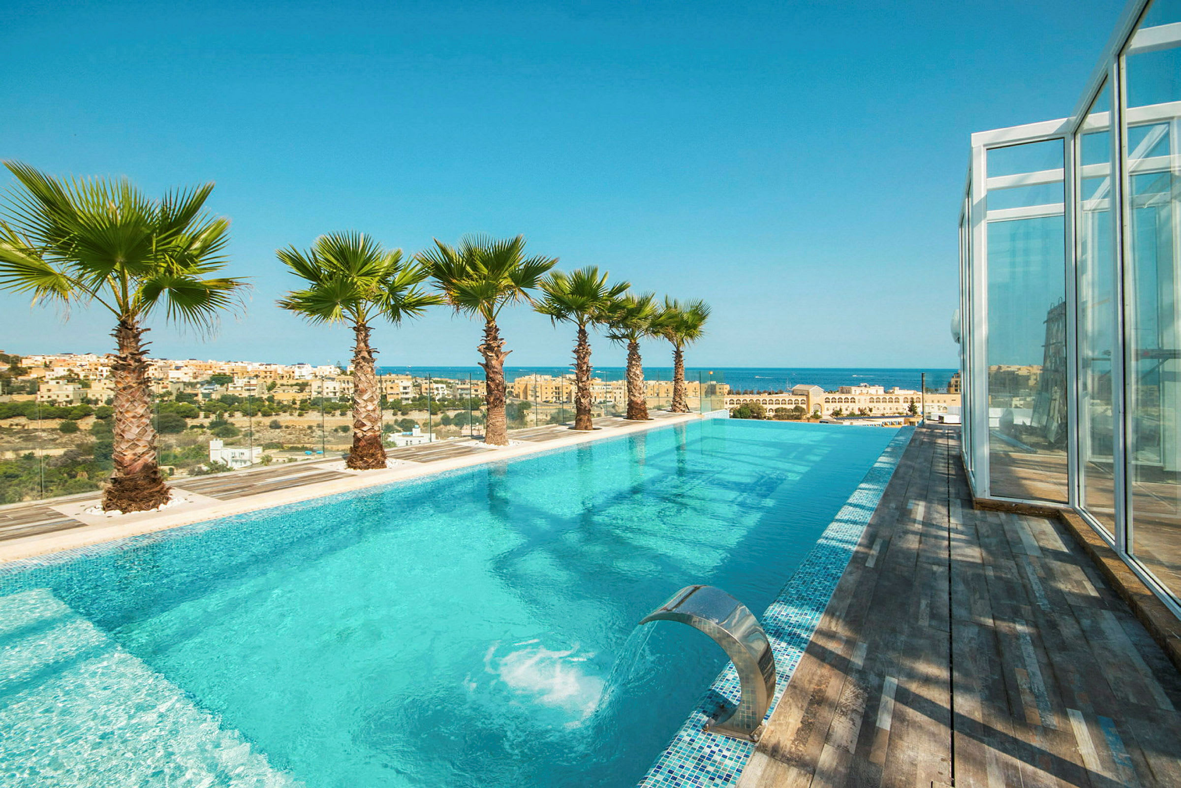 En infinitypool med utsikt över havet och staden, omgiven av palmer och trädeck. (AI-genererad text)