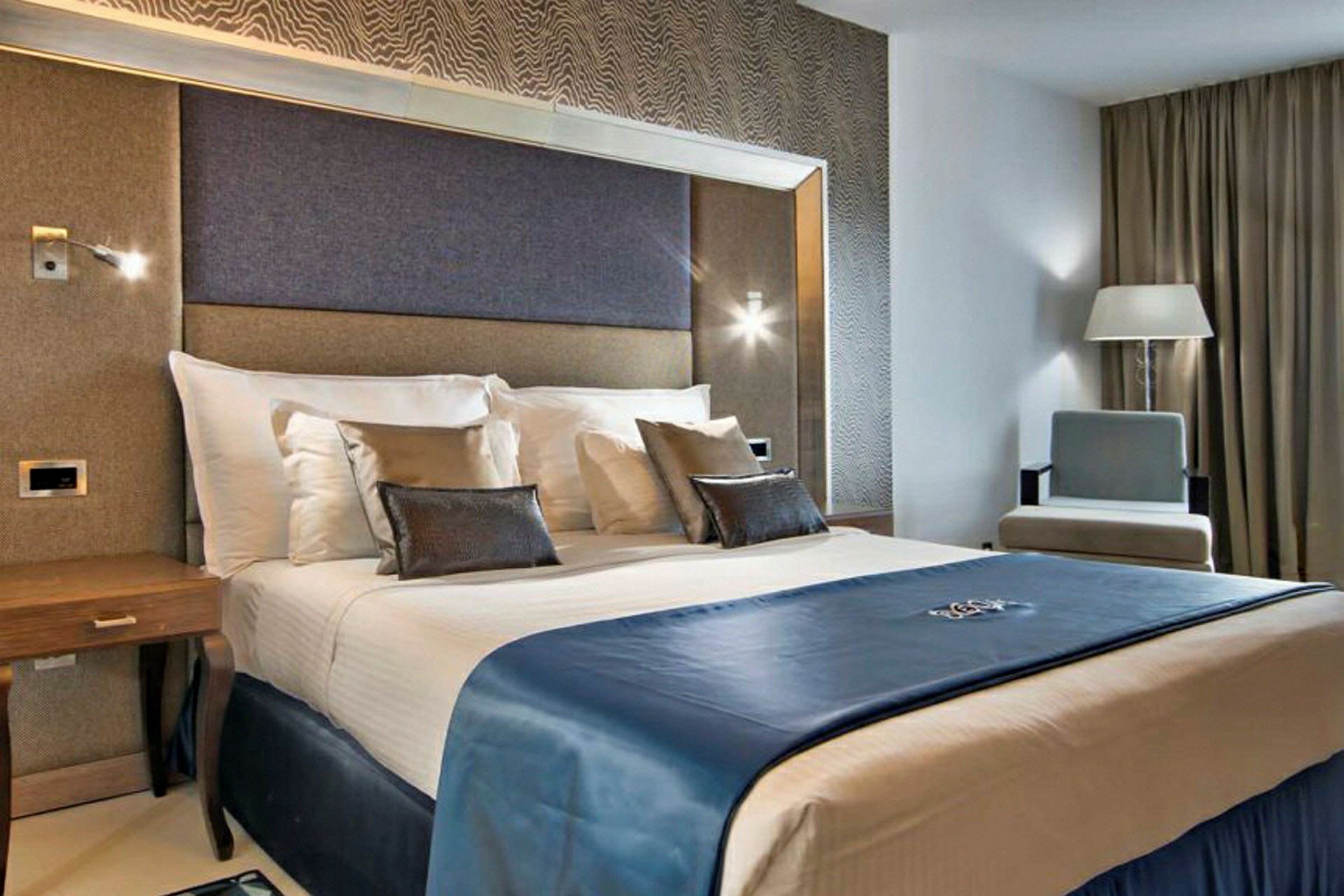 Ett hotellrum med modern inredning som visar en dubbelsäng med vita lakan och blå överkast, omgiven av eleganta möbler. (AI-genererad text)