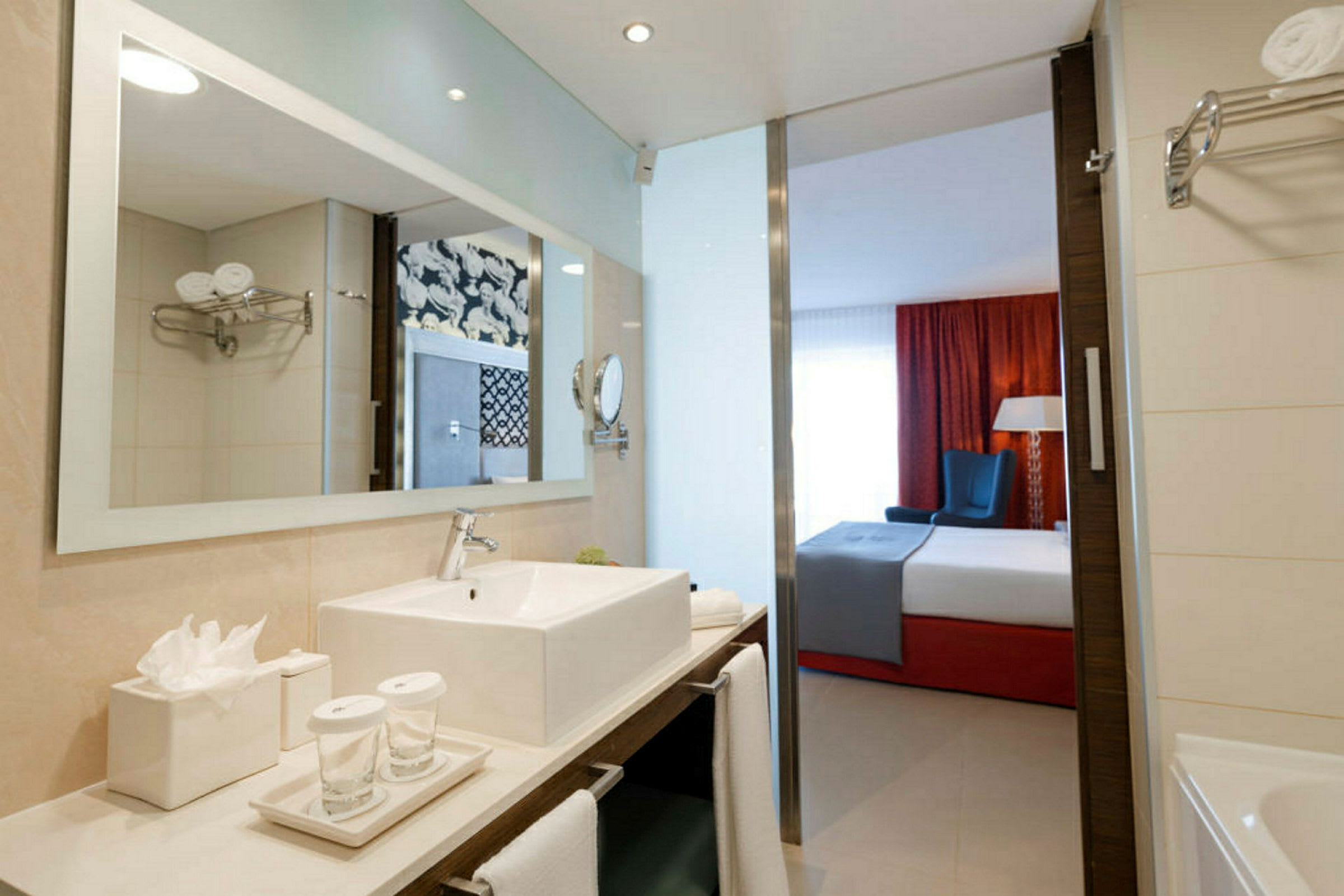 En modern hotelltoalett med vitt handfat och glas i förgrunden, med en vy genom en öppning till ett sovrum med säng och röd gardin. (AI-genererad text)