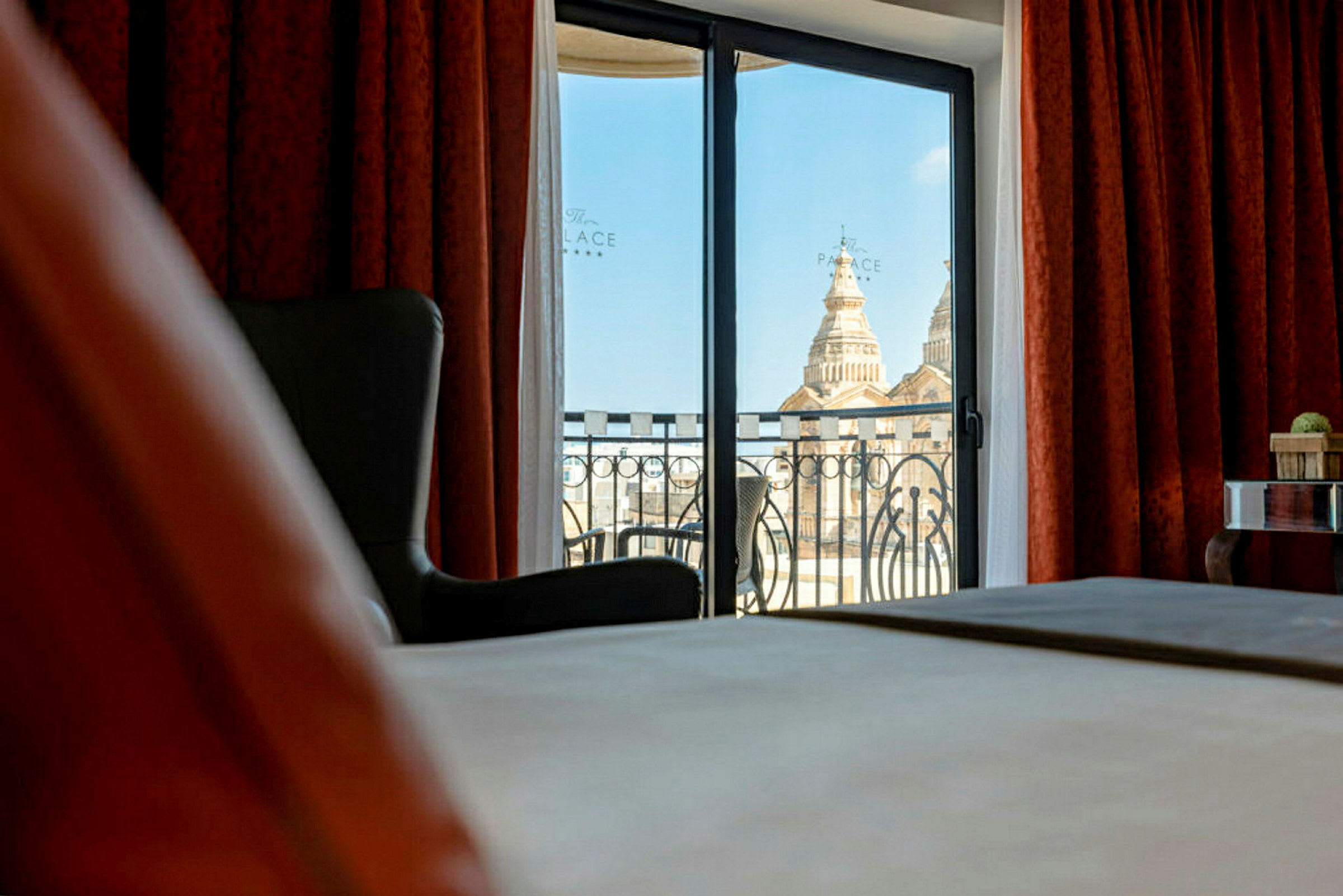 Ett öppet hotellfönster med utsikt över en kupol på avstånd, rödbruna gardiner och en balkongräcke. (AI-genererad text)
