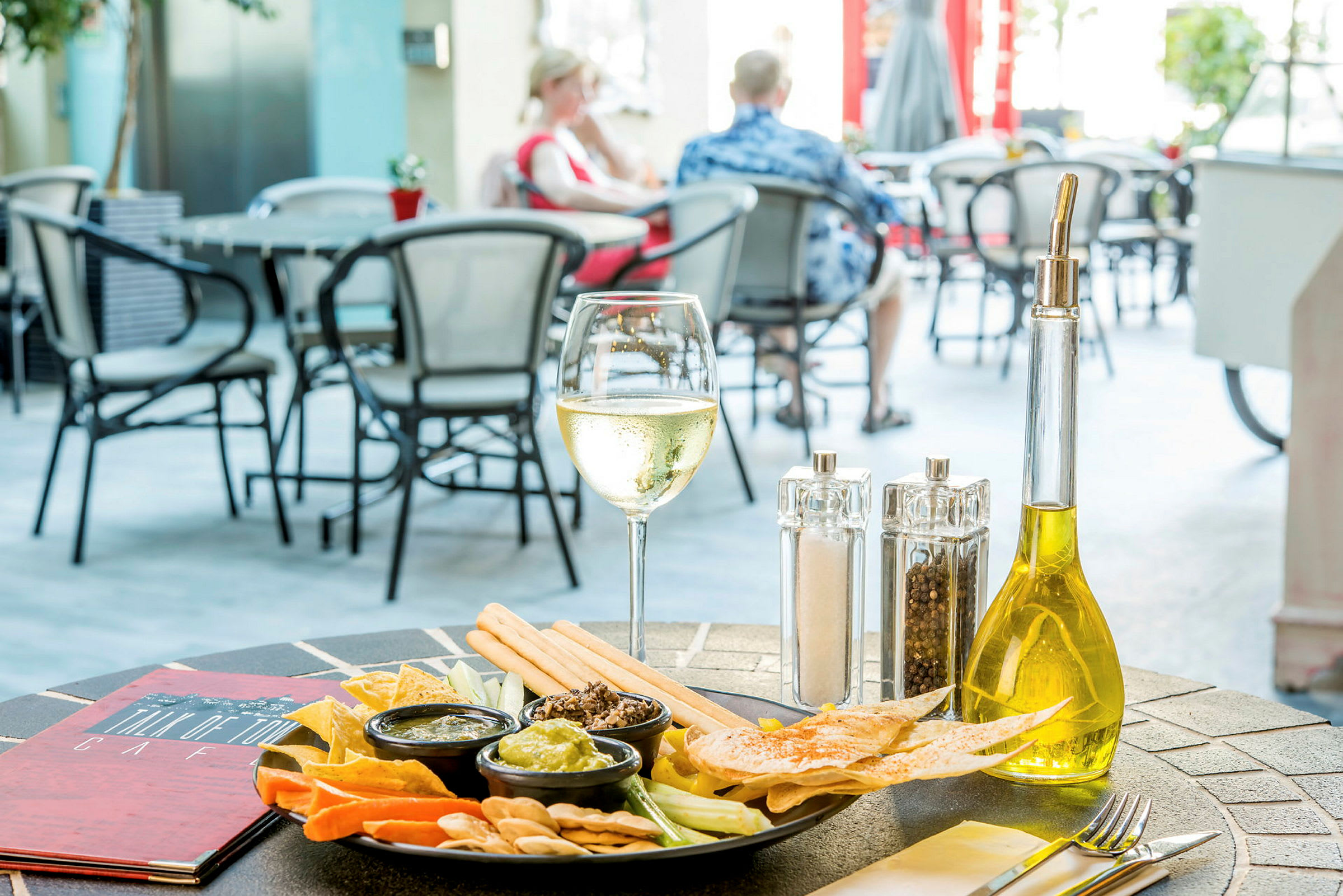 Ett bord på en utomhusrestaurang med en tallrik med snacks, små skålar med dipsås, ett glas vitt vin och en flaska olivolja. (AI-genererad text)