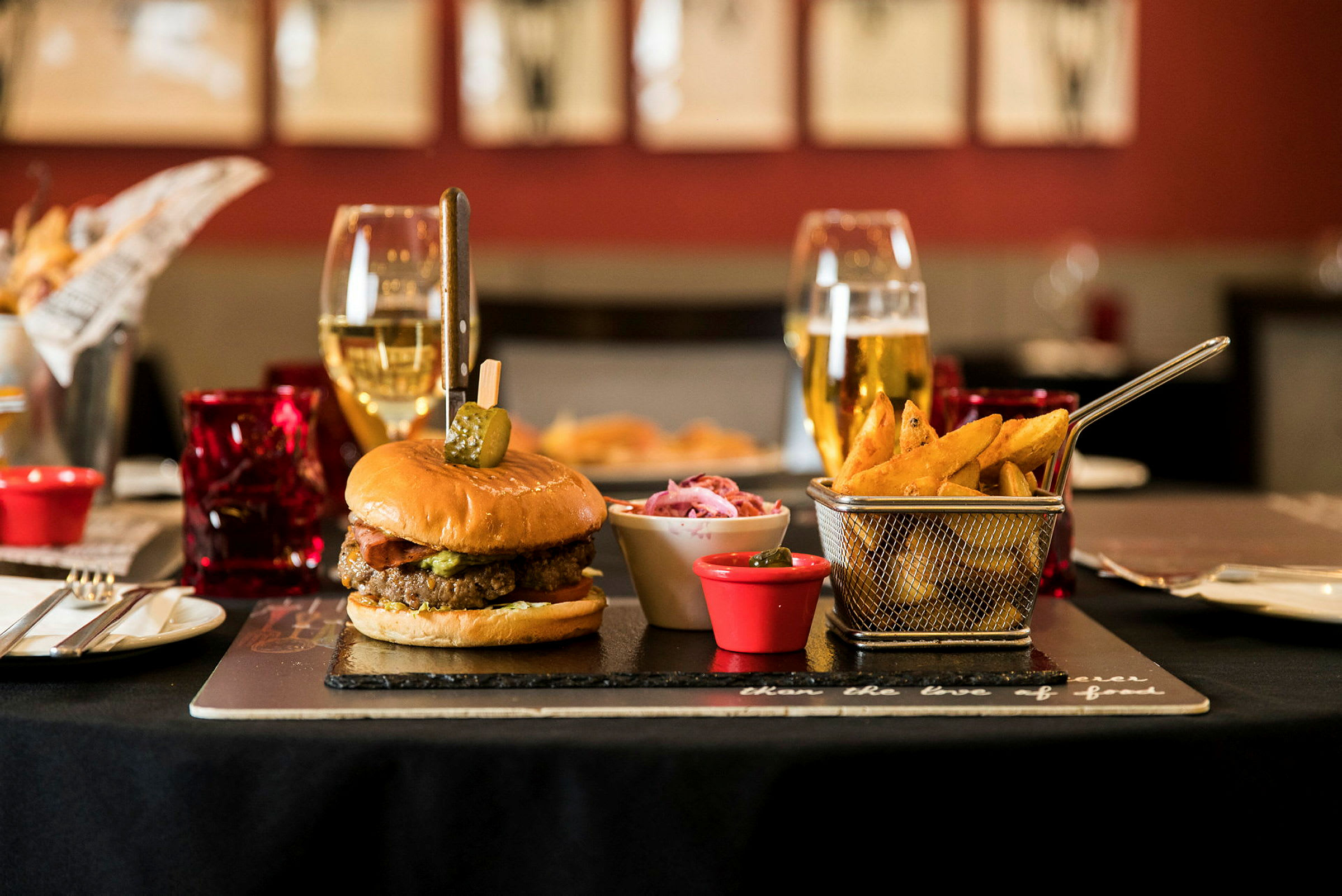 En hamburgare med pommes frites och tillbehör på ett bord, omgiven av glas och bestick. (AI-genererad text)