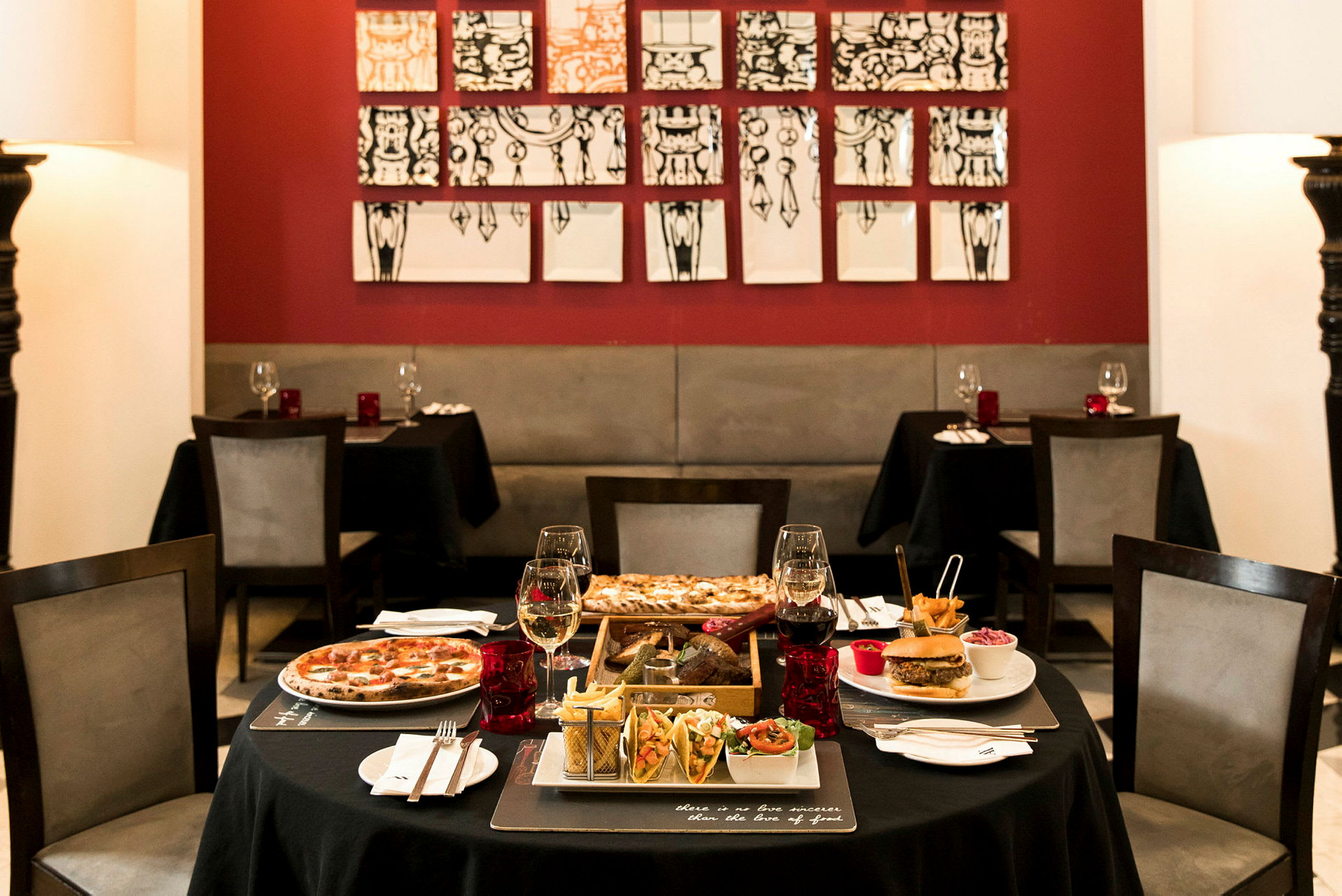Ett bord i en restaurang med olika maträtter och drycker, omgivande av röda väggar och modern konst. (AI-genererad text)
