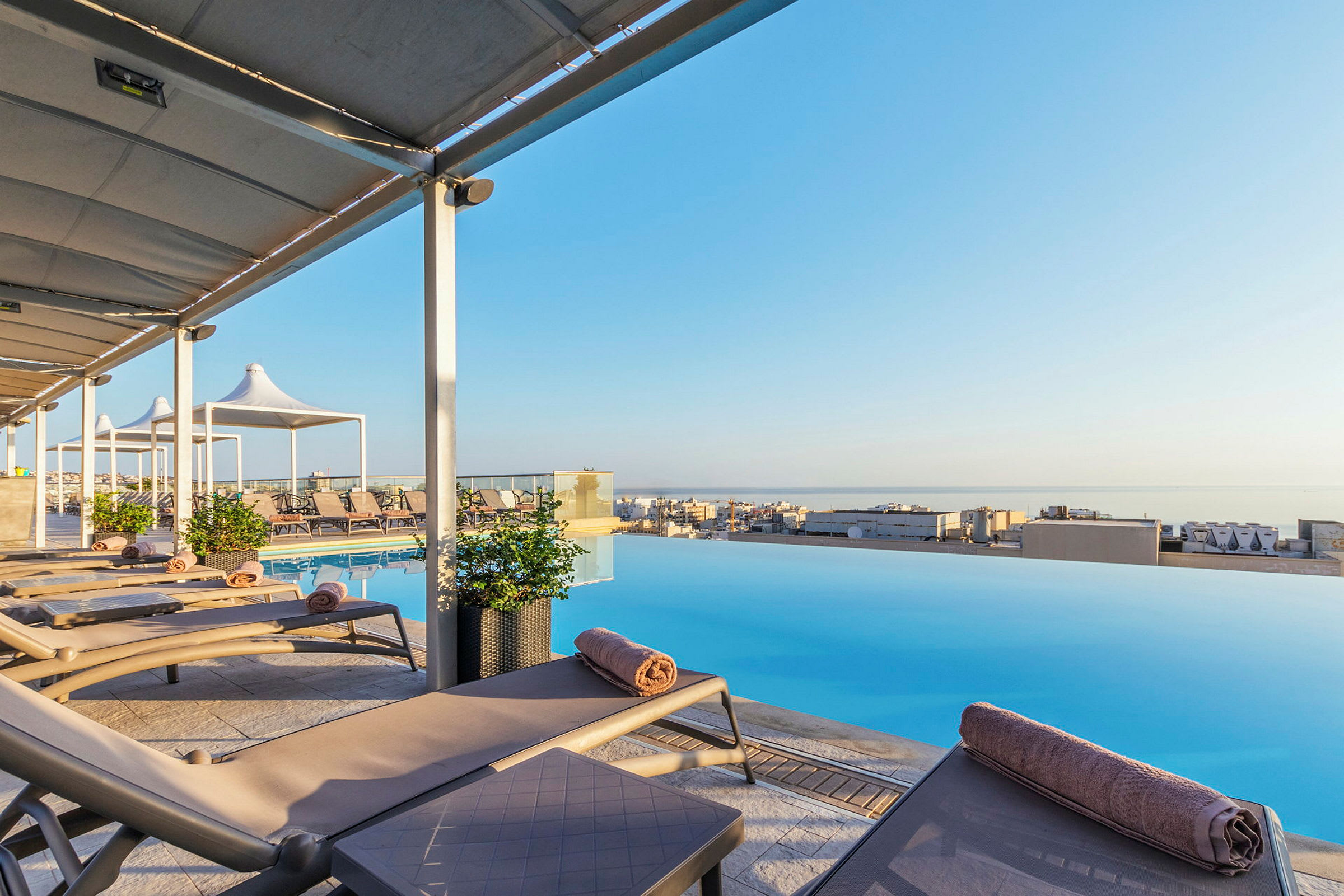 Solstolar vid en infinitypool med utsikt över en stad och havet. (AI-genererad text)