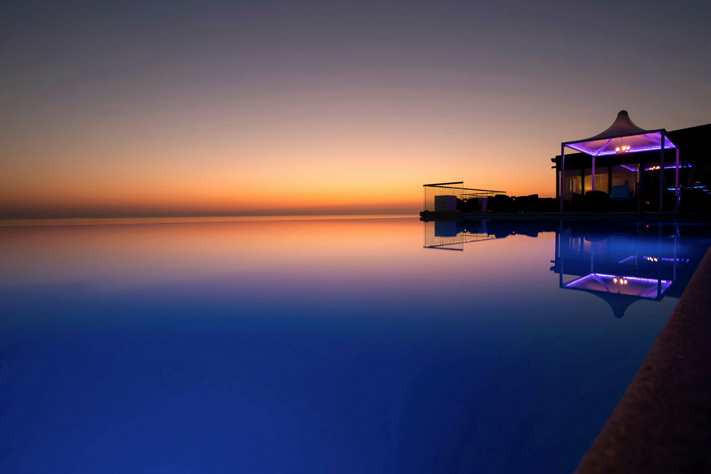 En infinitypool med utsikt över en solnedgång. Poolen reflekterar den färgglada himlen och ett upplyst område vid kanten. (AI-genererad text)