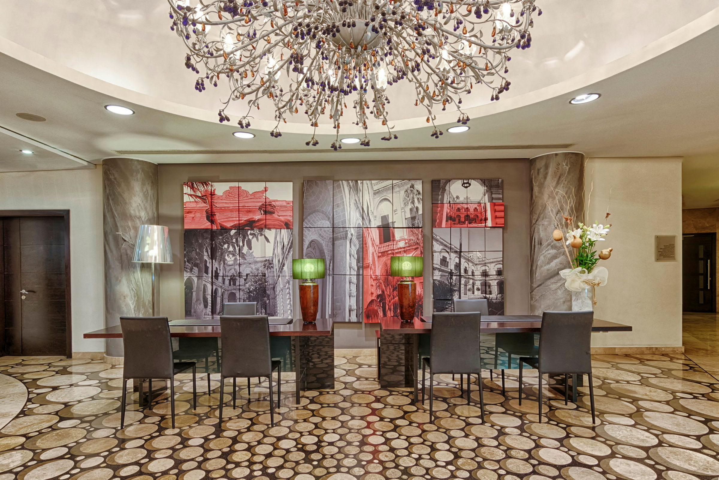 Ett elegant hotellobby med ett stort dekorativt taklamp som hänger över flera skrivbord med stolar, målningar på väggen bakom, och vackra dekorationer som blommor och bordslampor. (AI-genererad text)