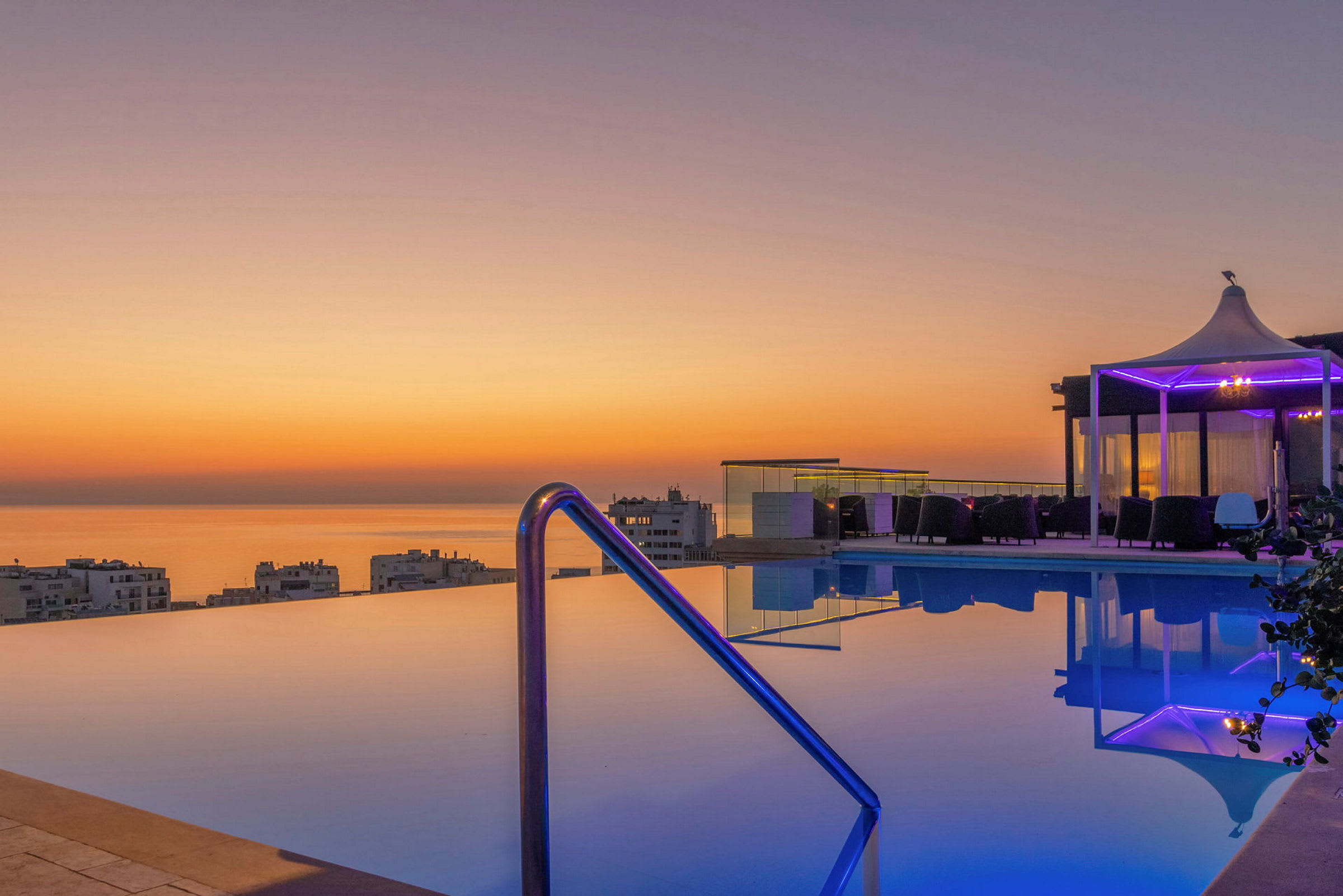 En infinitypool med utsikt över havet och staden, omgiven av solnedgångens varma färger. (AI-genererad text)