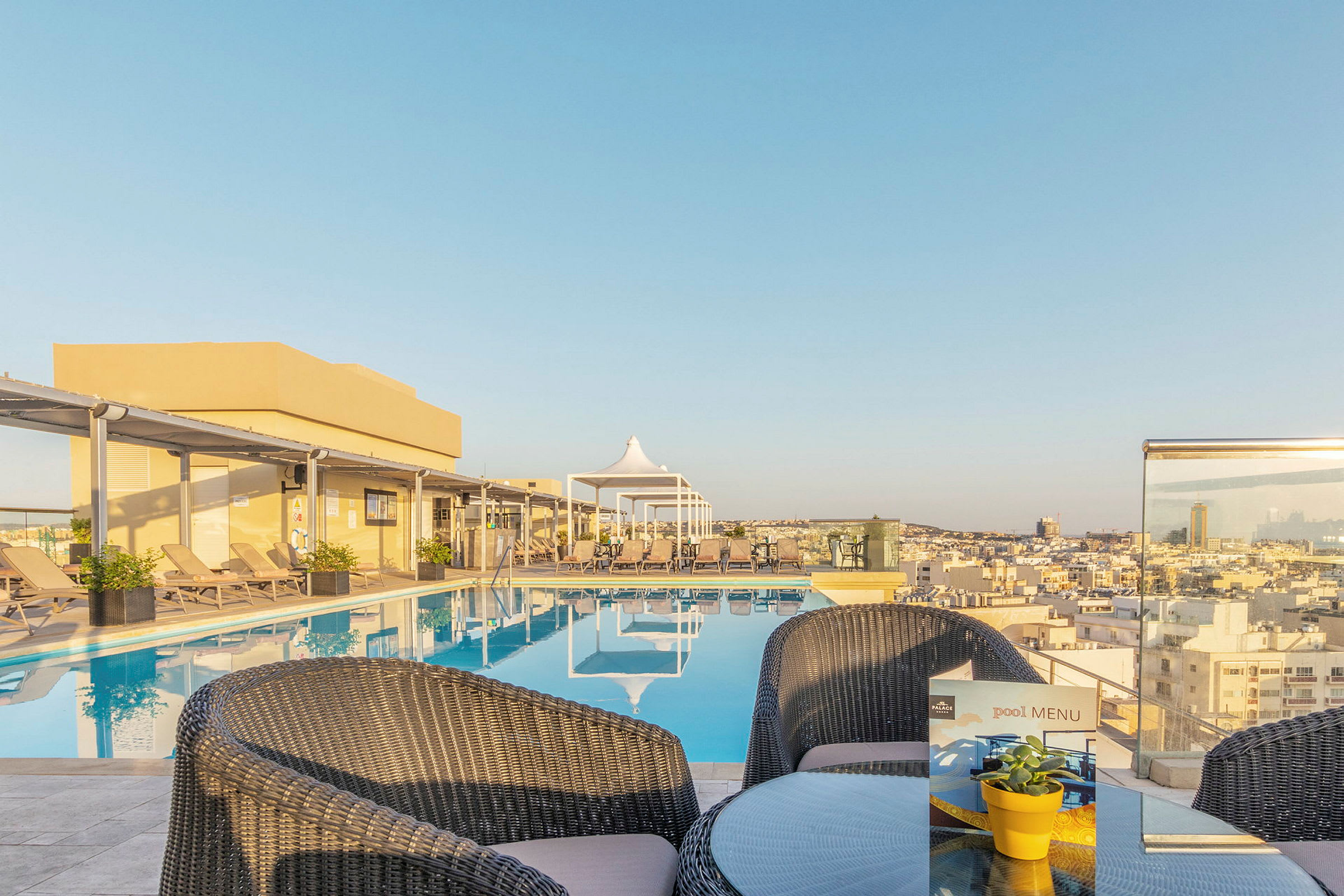 Bild på en pool på en hotellterrass med utsikt över staden, omgiven av solstolar och ett mindre bord med stolar. (AI-genererad text)