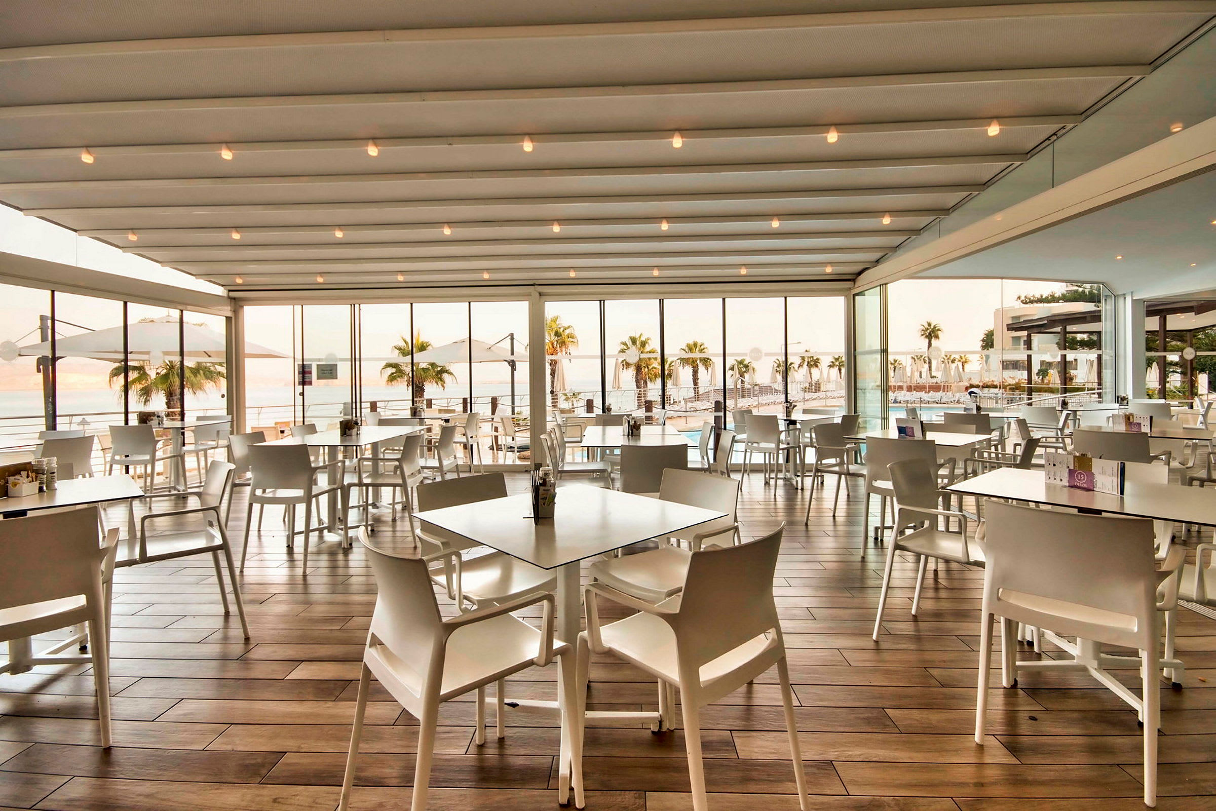 En restaurang med vit inredning, stora fönster och utsikt över en pool och palmer utanför. (AI-genererad text)
