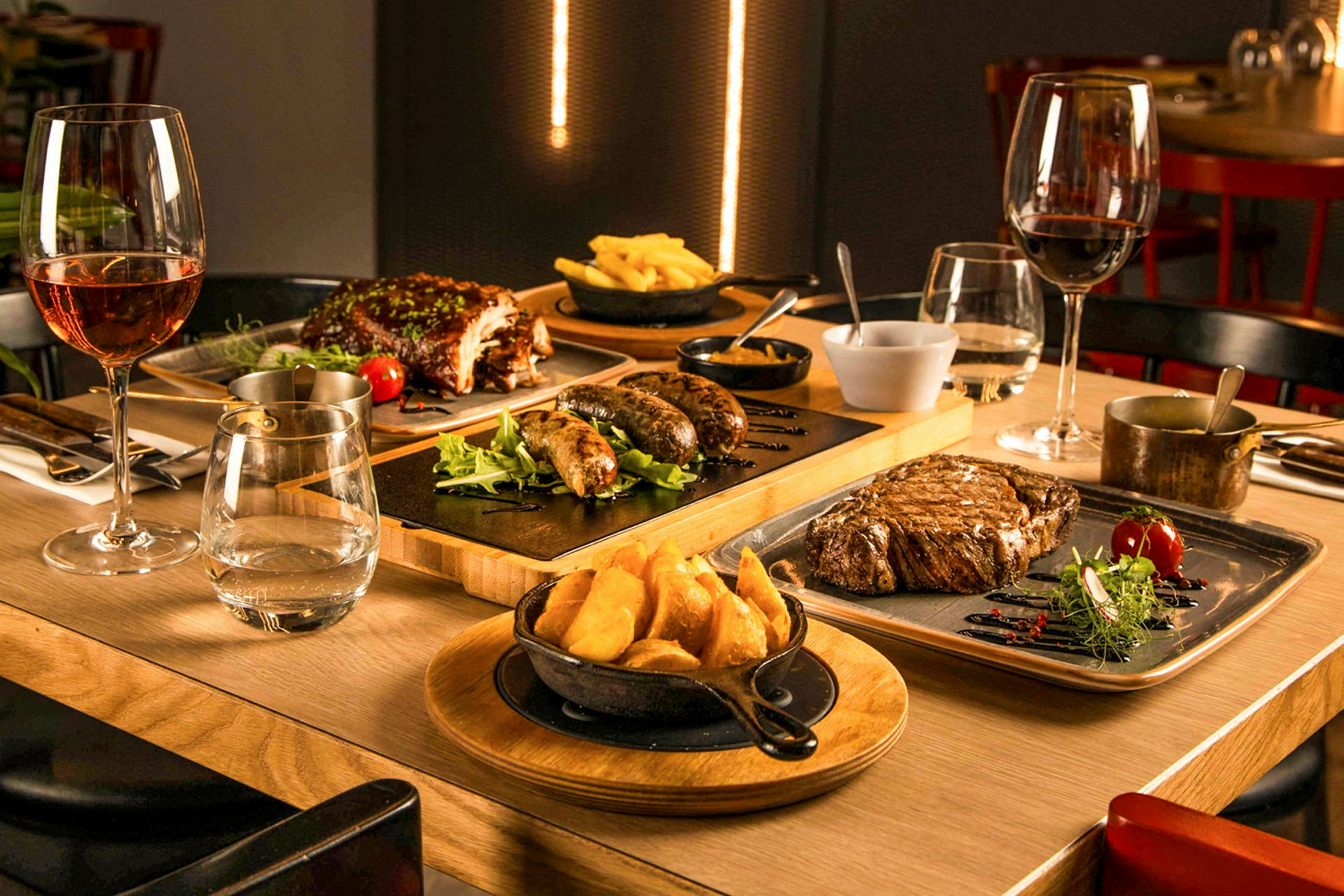 Ett bord är dukat med olika maträtter, däribland stek, korvar, ribs och pommes frites, tillsammans med vinglas. (AI-genererad text)