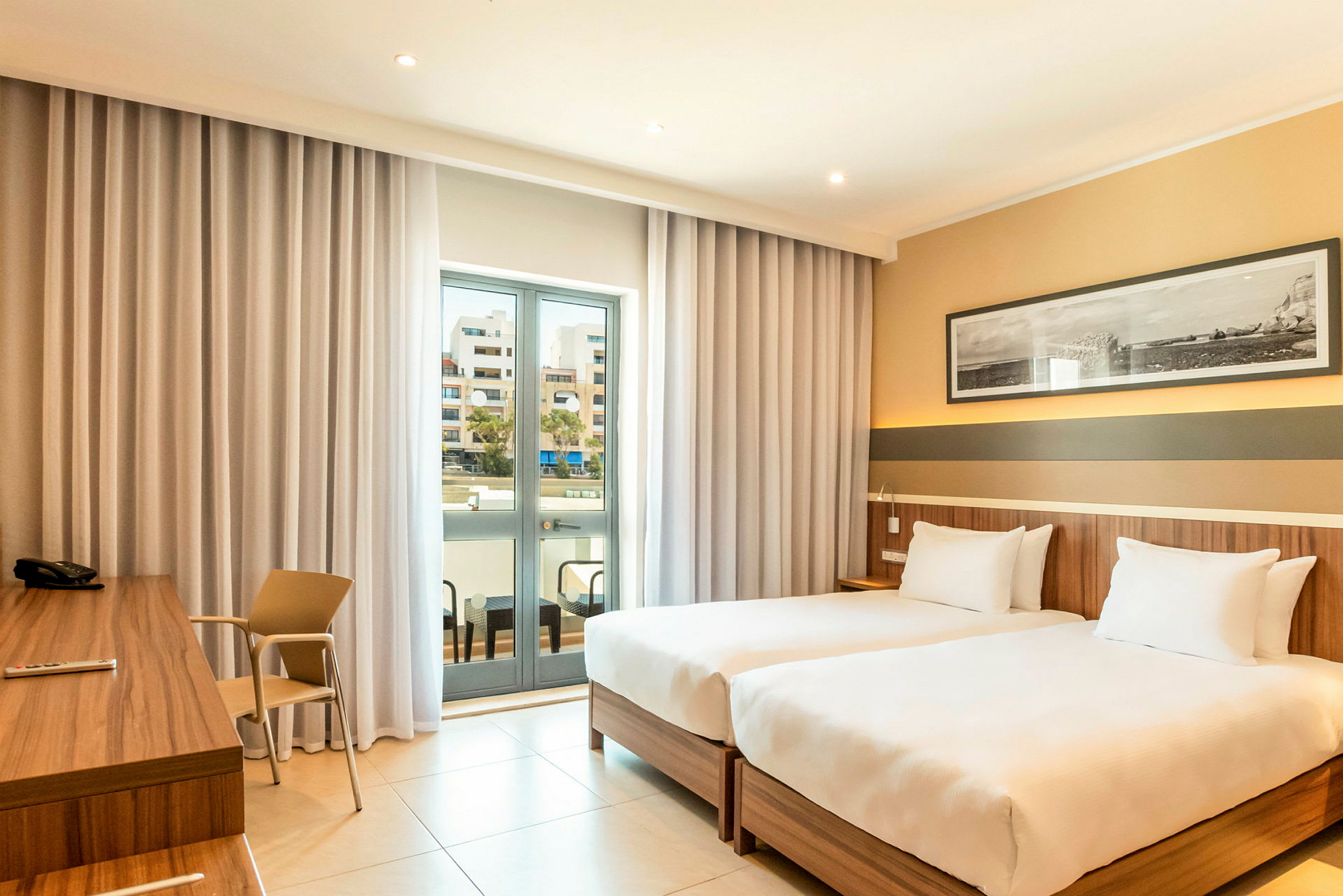 Hotellrum med två enkelsängar, skrivbord och en balkong med glasdörrar. (AI-genererad text)