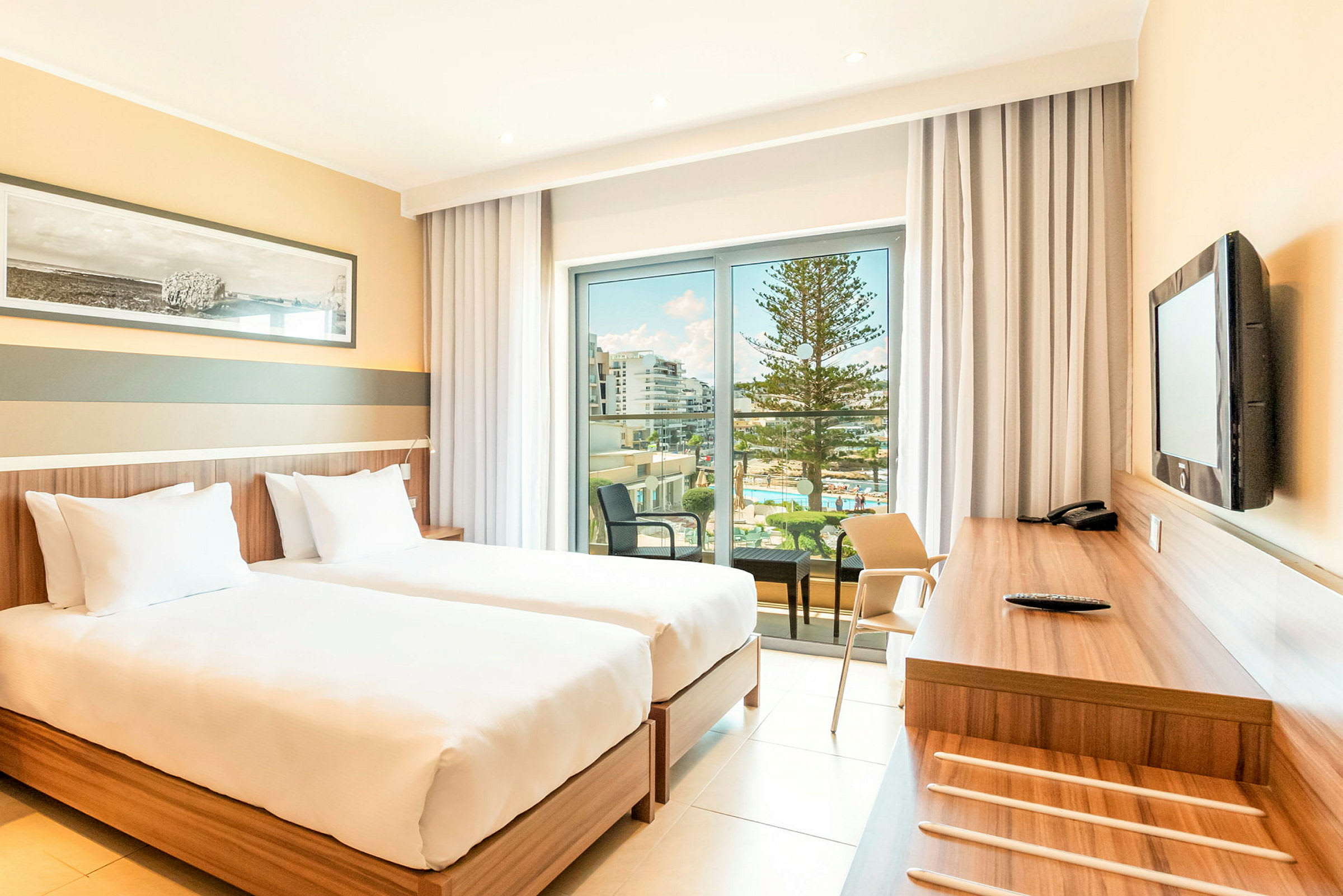 Ett hotellrum med två enkelsängar, ett skrivbord och utsikt över en balkong med pool och träd. (AI-genererad text)