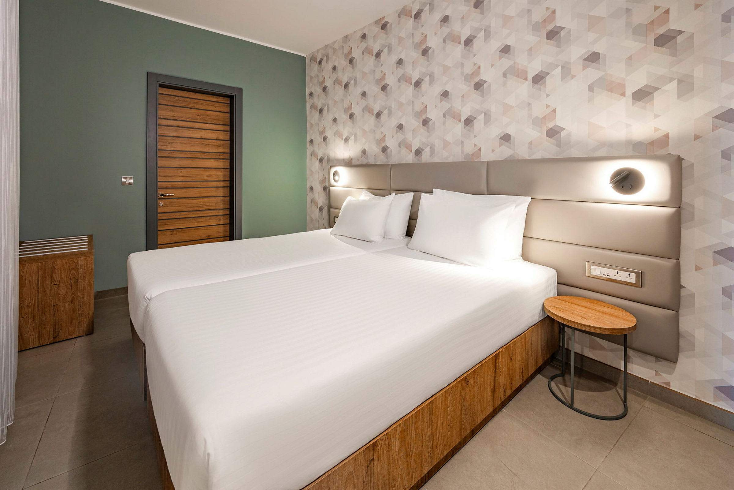 Ett modernt hotellrum med två enkelsängar som står tillsammans, en träpaneldörr och geometriskt mönstrad tapet. (AI-genererad text)