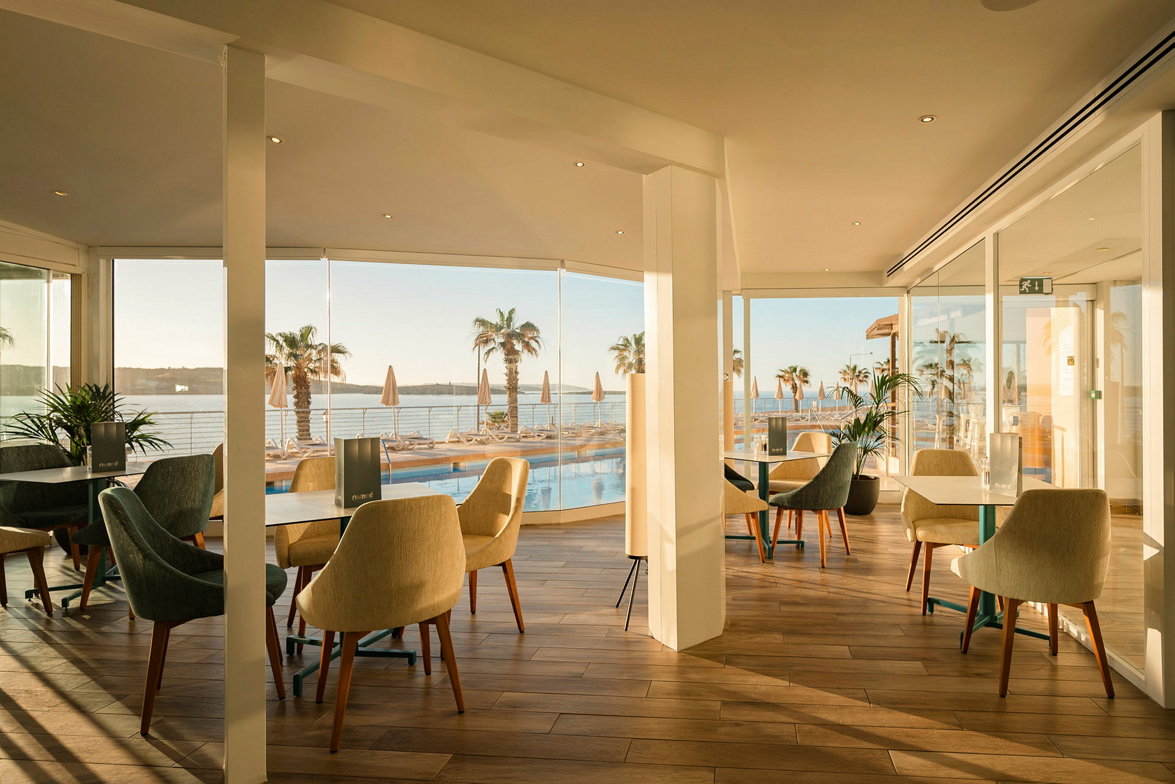En restaurang med sittplatser och glasväggar med utsikt över en pool och palmer vid havet. (AI-genererad text)