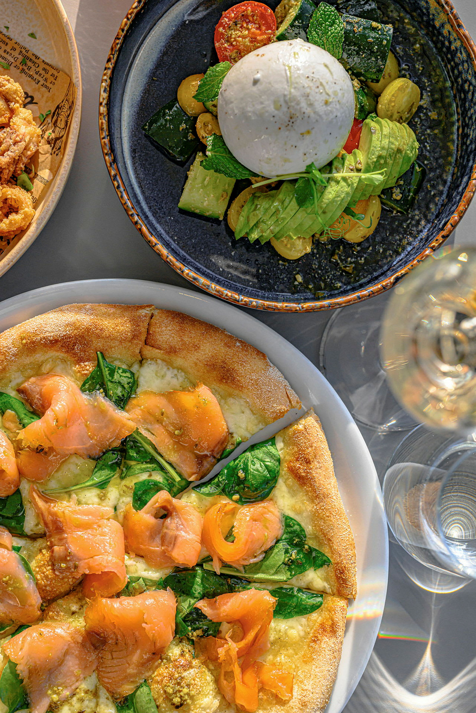 En tallrik med burrata, grönsaker och avokado, samt en pizza med lax och spenat. (AI-genererad text)