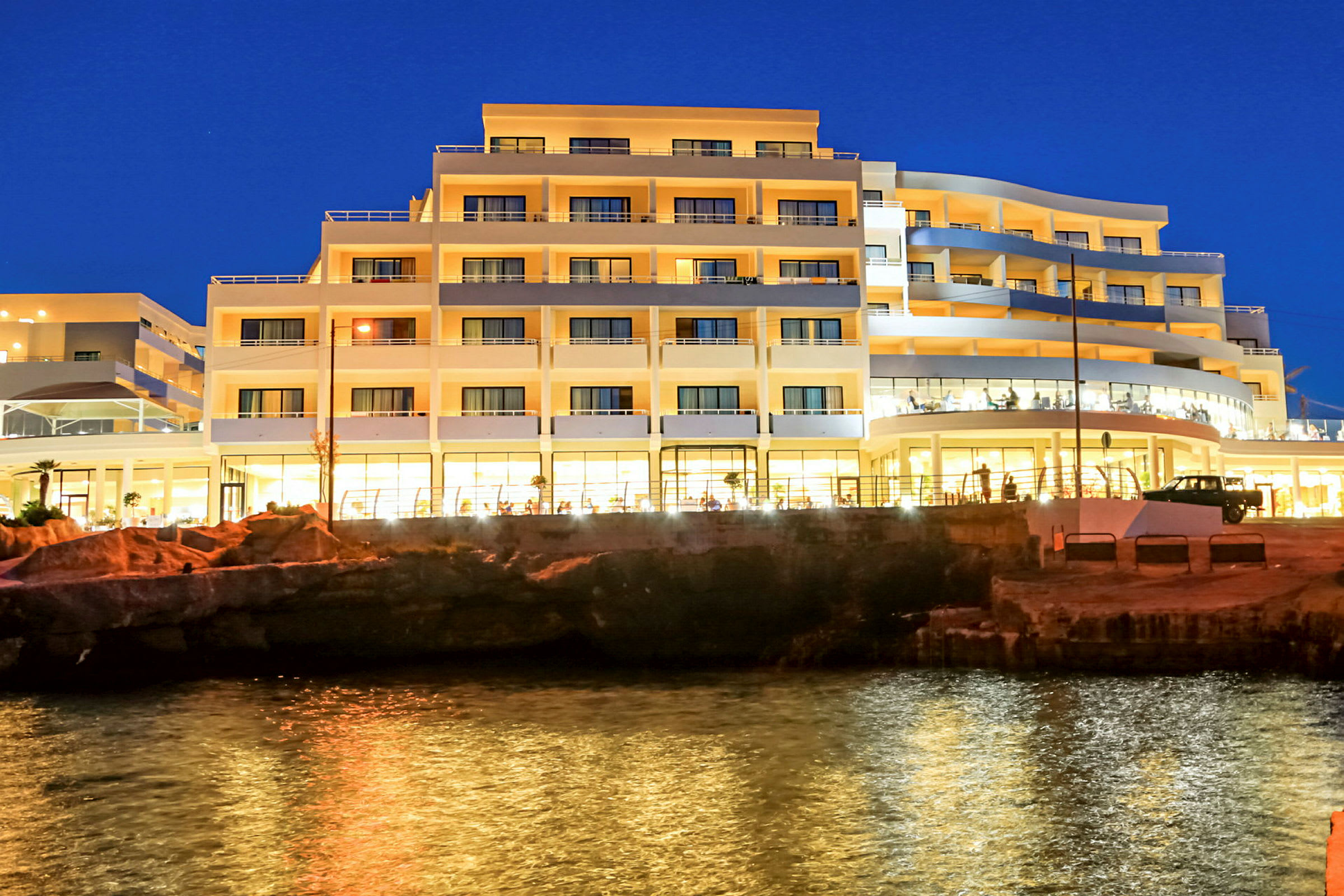 En hotellbyggnad vid en kust på kvällen, med ljus som speglas i vattnet. (AI-genererad text)