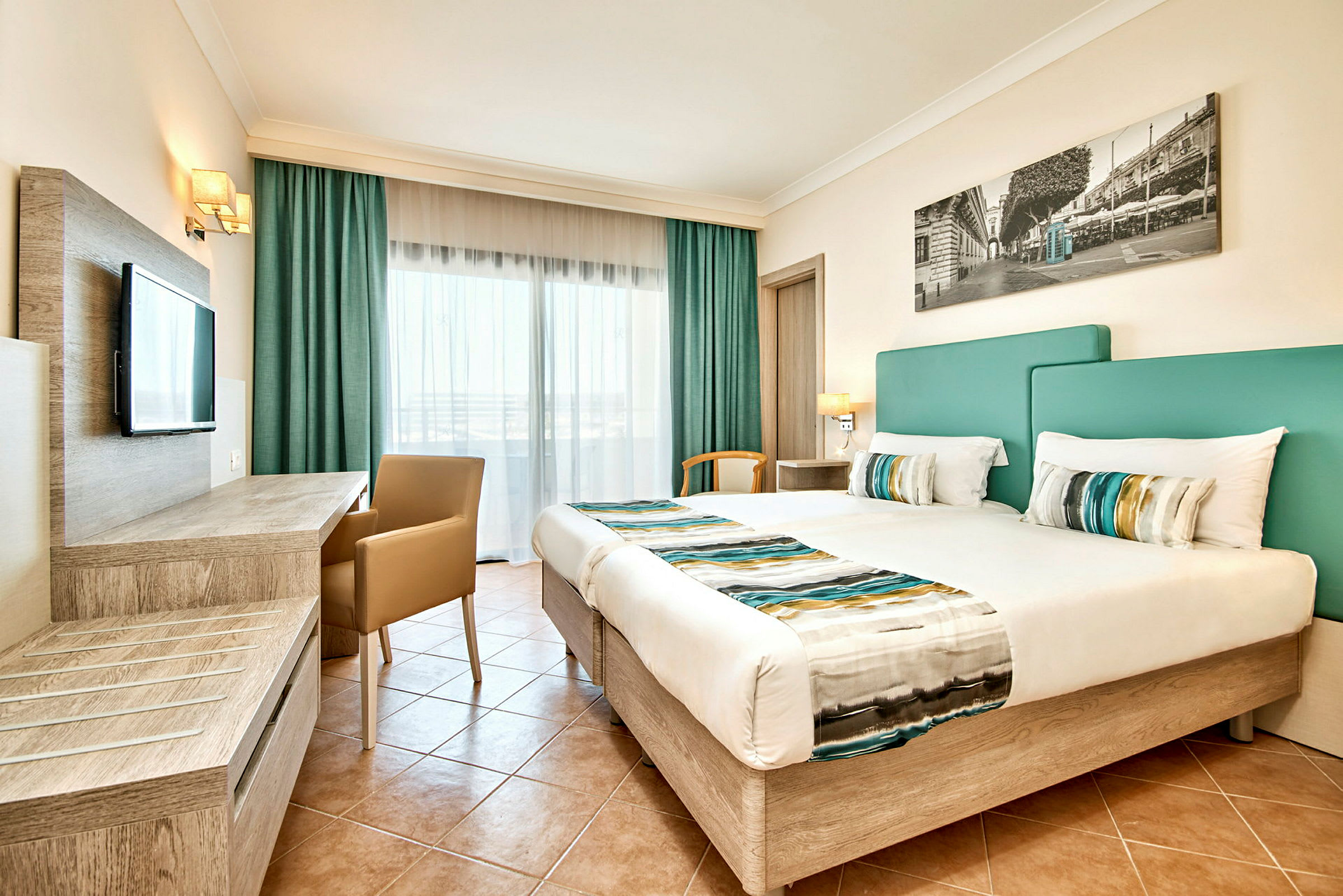 Ett ljust hotellrum med två sängar, grönklädda sänggavlar och färgglada sängdukar, med ett skrivbord och en tv på väggen. (AI-genererad text)