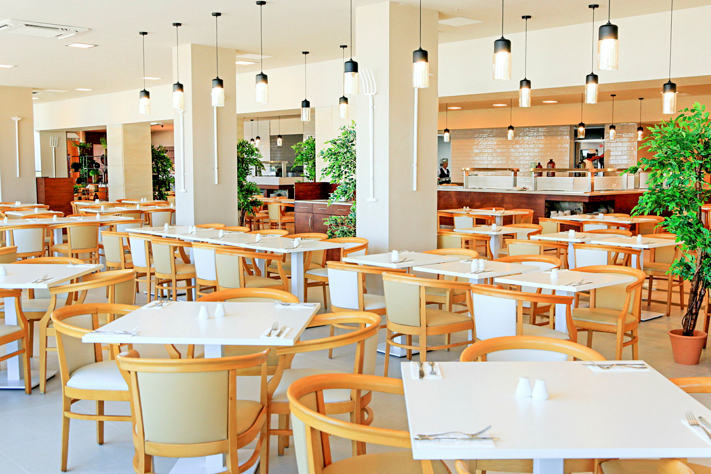 En restaurang med moderna vita bord och ljusa trästolar, dekorerad med hängande lampor och växter. (AI-genererad text)