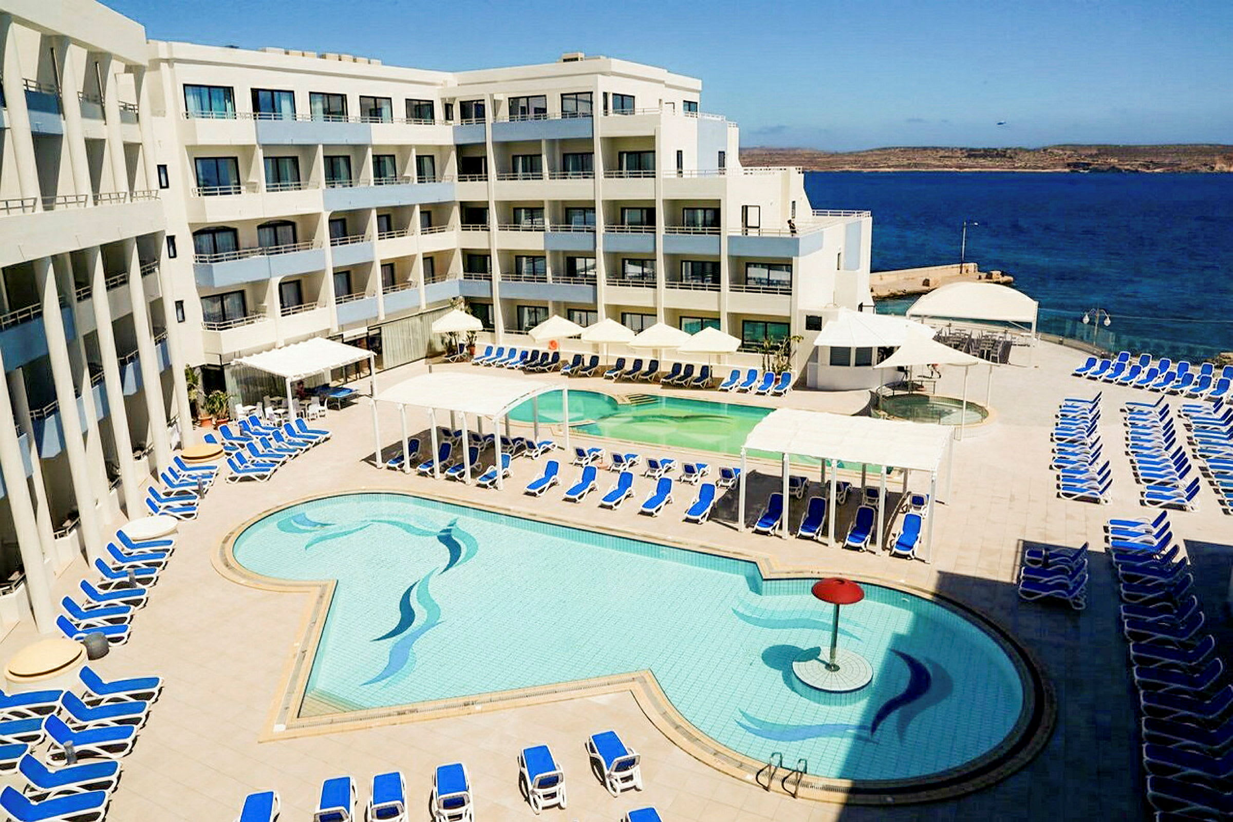 En hotellpool med solstolar och havsutsikt i bakgrunden. (AI-genererad text)