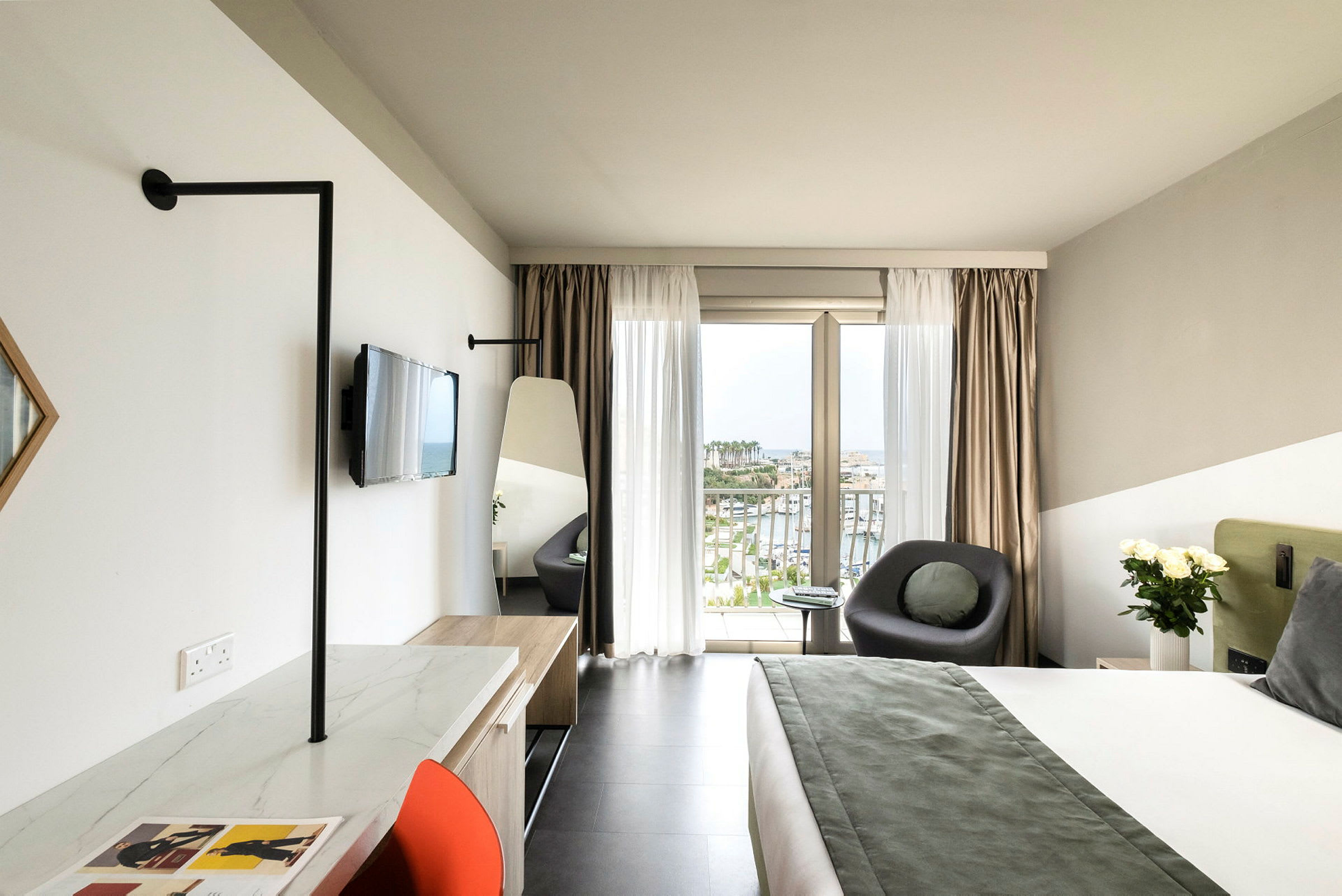 Ett modernt hotellrum med en stor säng, en sittgrupp vid ett fönster med utsikt över en hamn, samt en skrivbordsplats med en stol. (AI-genererad text)