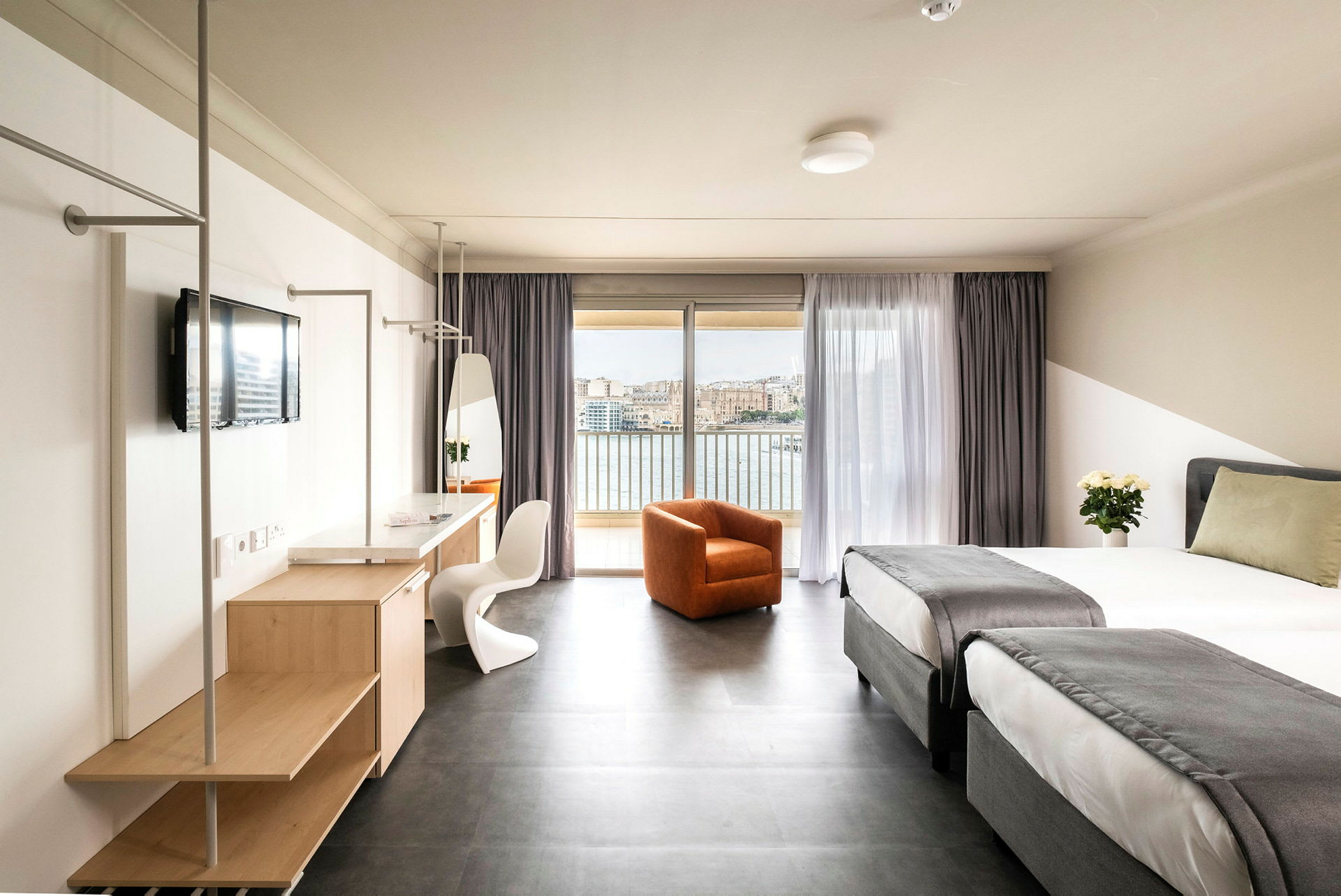 Ett hotellrum med två enkelsängar med grå överkast, en orange fåtölj, ett skrivbord med spegel och tv samt en balkong med utsikt över vatten och en stad. (AI-genererad text)
