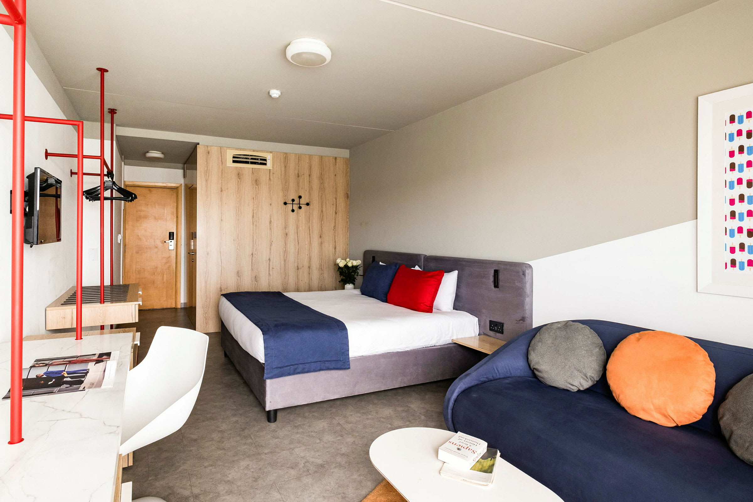 Ett hotellrum med en säng med blått överkast och röda och vita kuddar, en blå soffa med färgglada kuddar, samt ett träbord och garderob. (AI-genererad text)