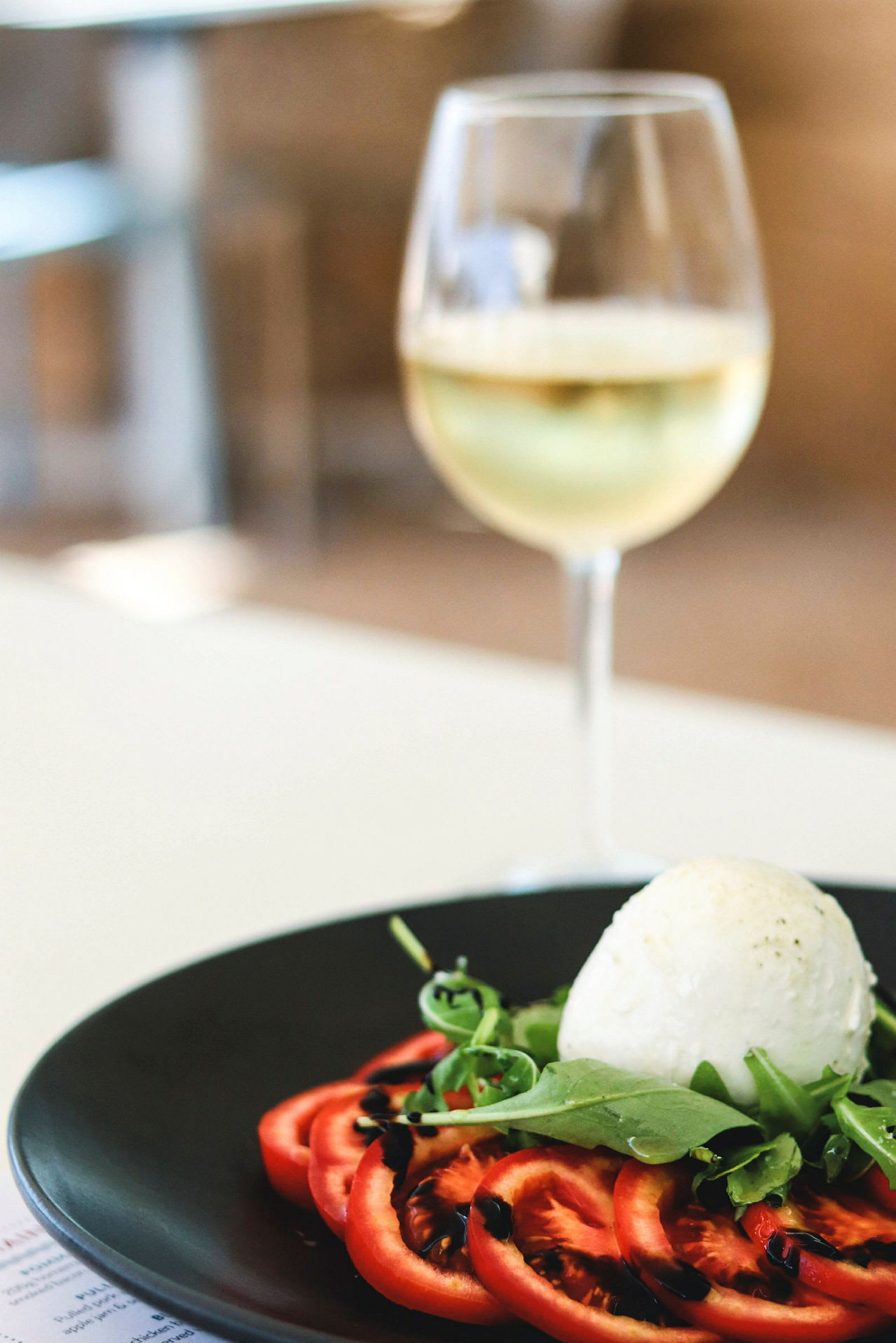 En tallrik med skivade tomater, ruccola och en boll av mozzarella, toppad med balsamicokräm. I bakgrunden ett glas vitt vin. (AI-genererad text)