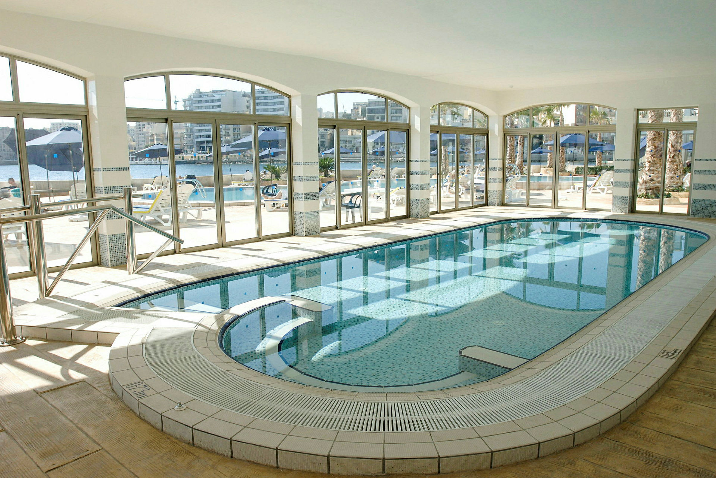 En inomhuspool med glasade väggar som visar utsikten mot en terrass med sittmöbler och parasoller. (AI-genererad text)