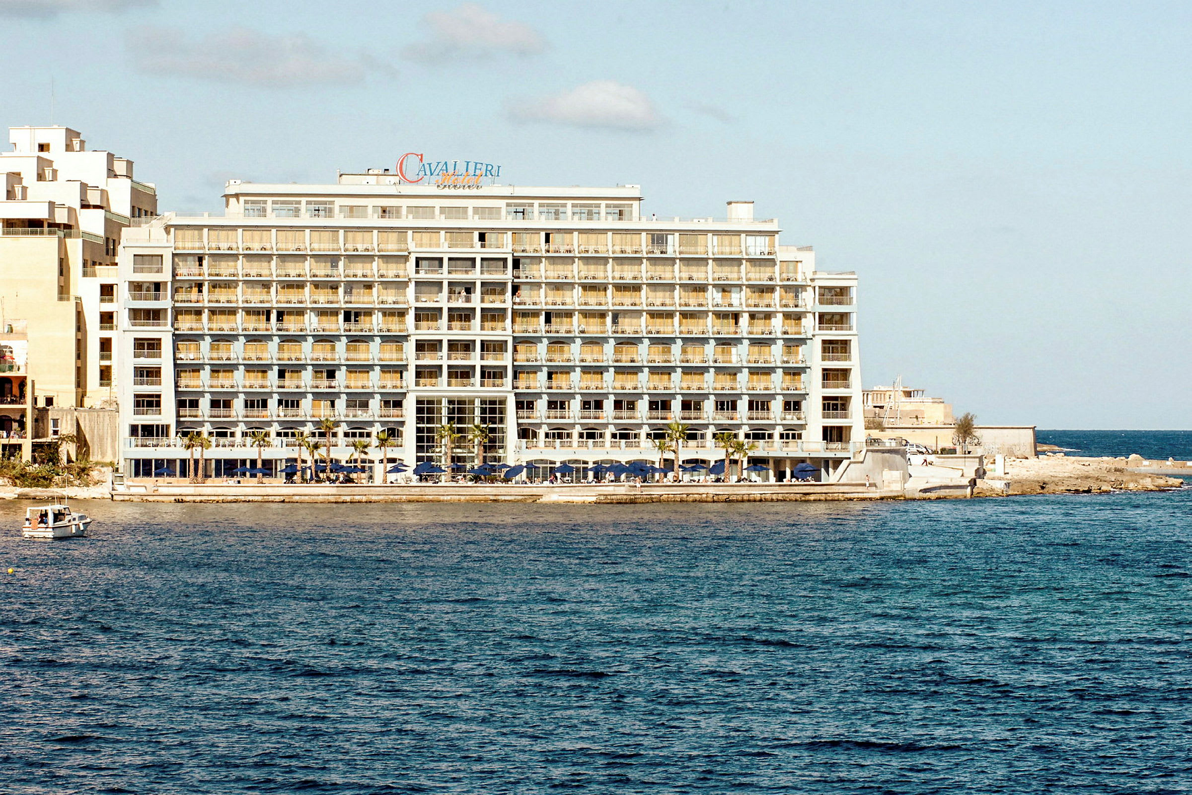 Ett stort hotell vid havet, med flera våningar och balkonger samt en liten båt i vattnet nära hotellet. (AI-genererad text)
