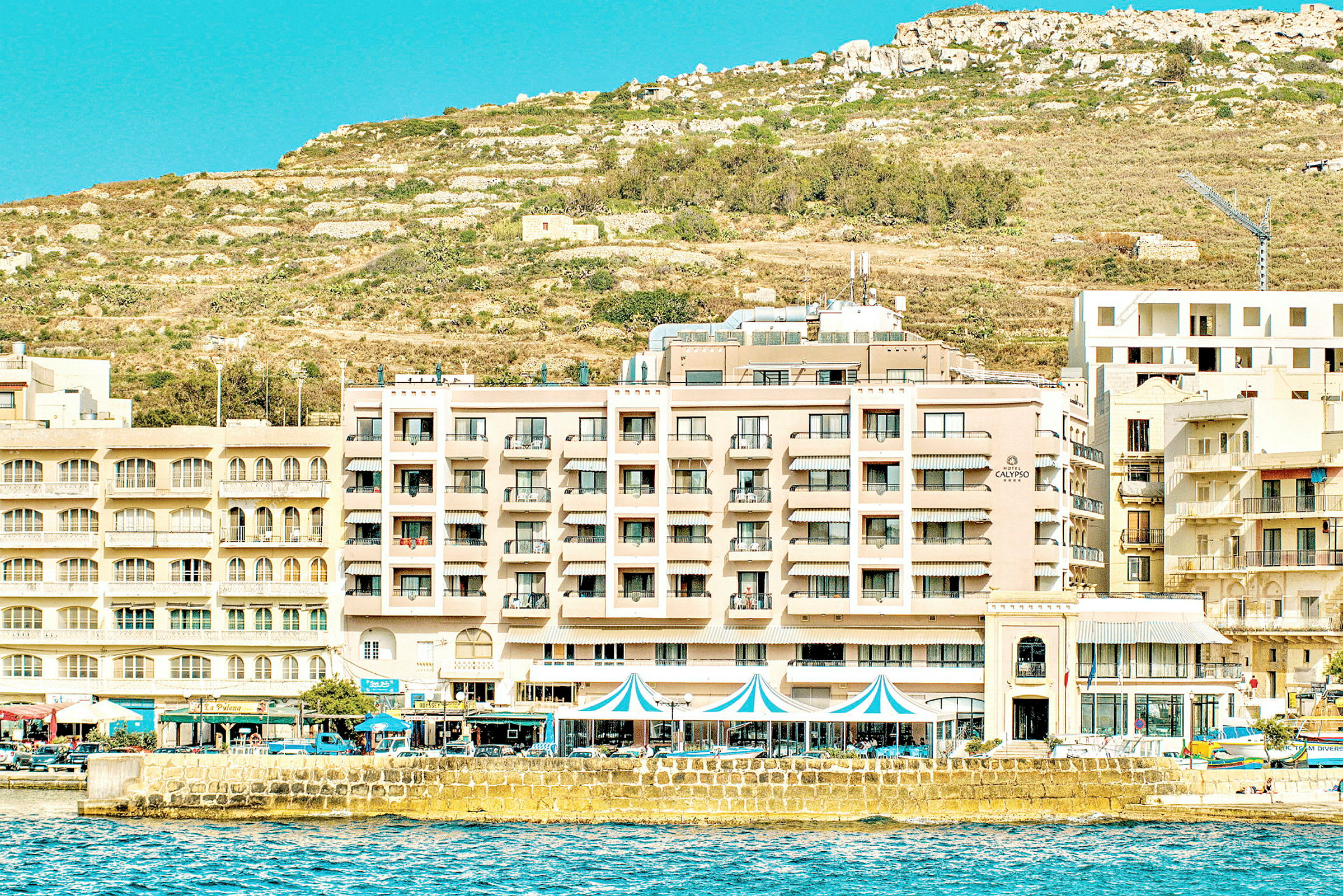 Hotellfasad vid strandpromenaden med kullig terräng i bakgrunden. (AI-genererad text)