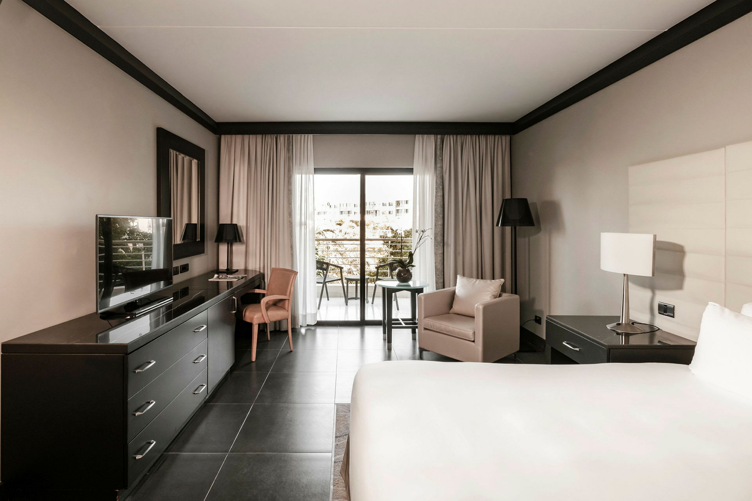 Hotellrum med modern inredning, en stor säng, skrivbord med TV, fåtölj och fönster mot en balkong. (AI-genererad text)