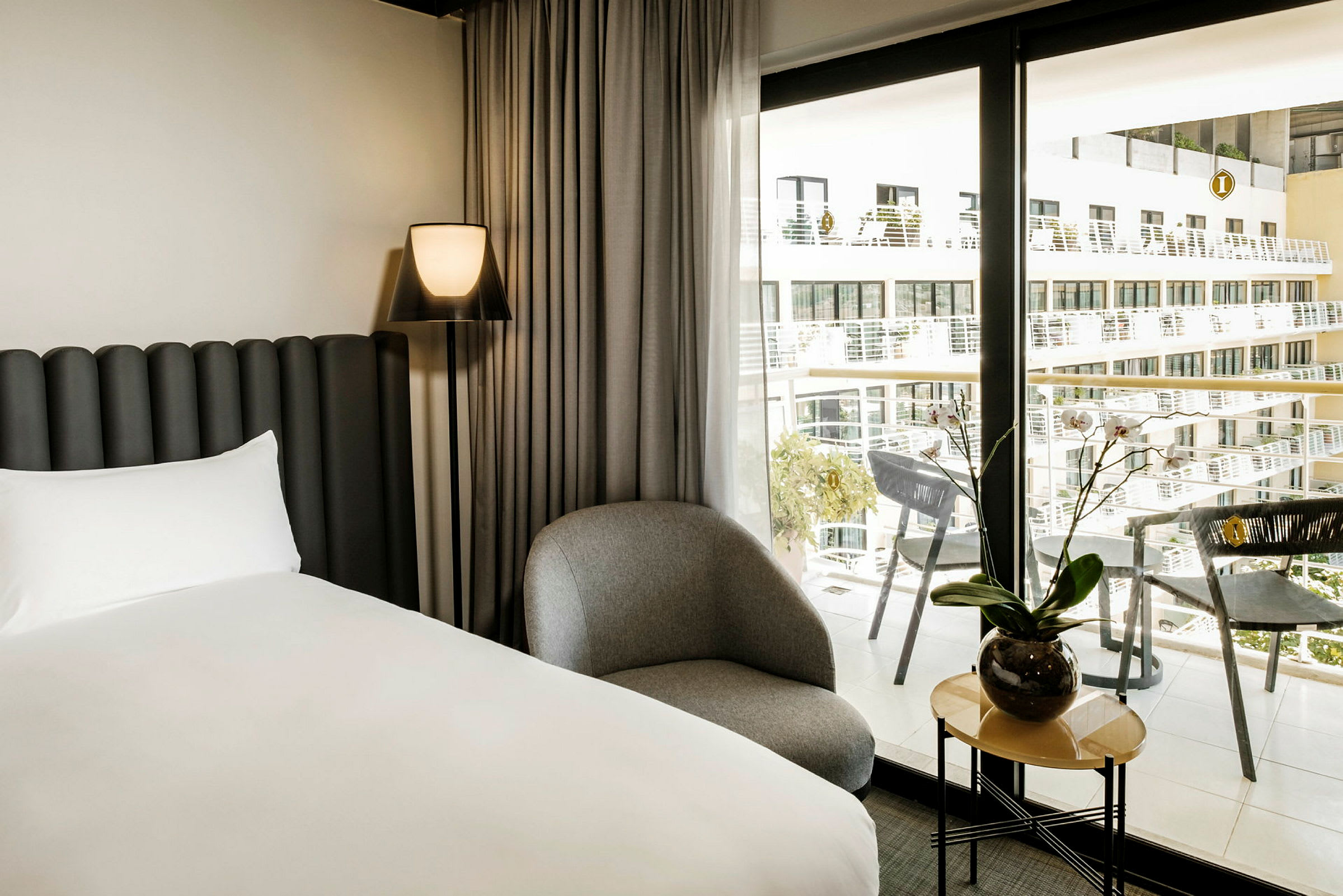 Ett modernt hotellrum med en säng, en stol och ett litet bord med en växt bredvid ett stort fönster med utsikt över balkonger. (AI-genererad text)