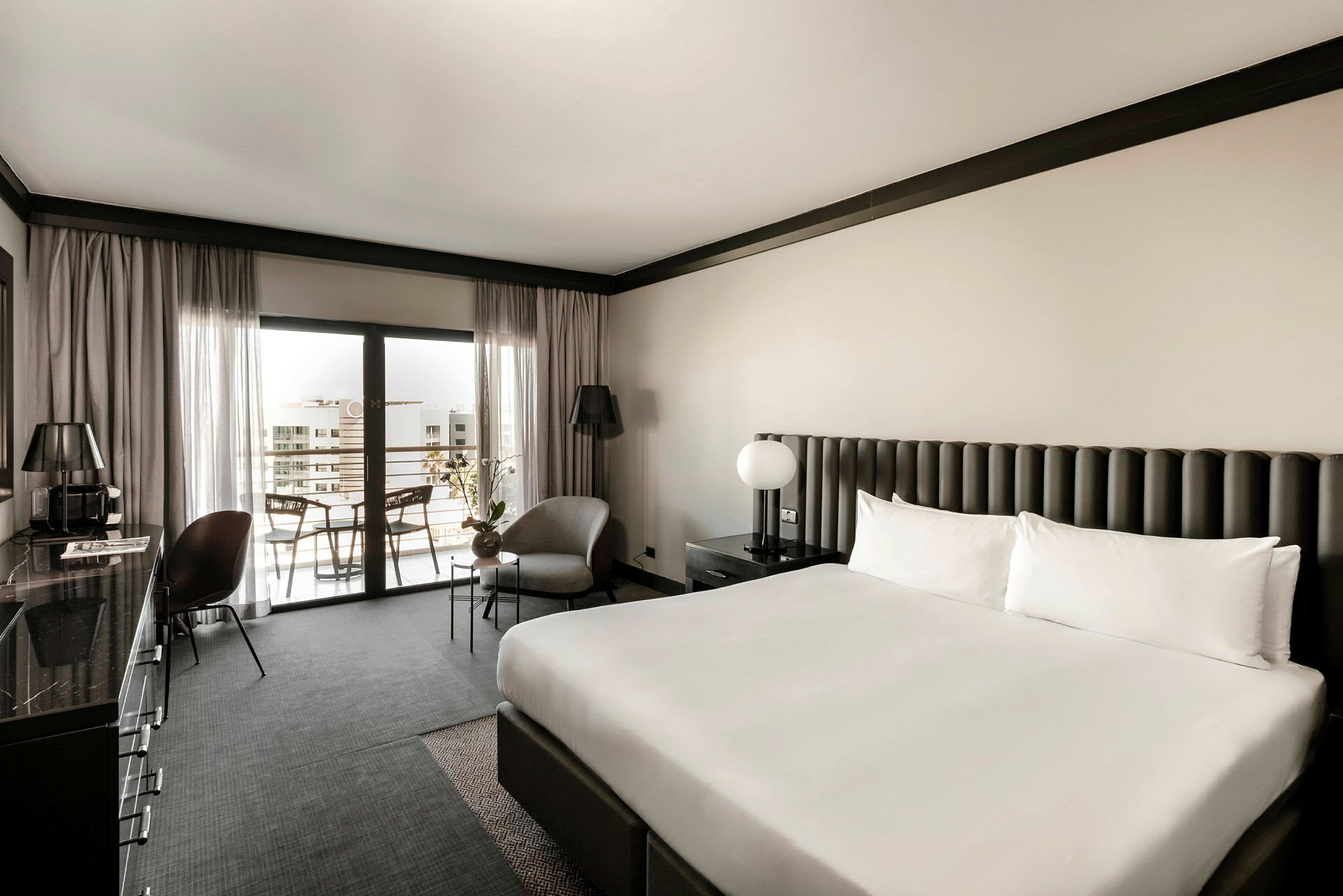 Hotellrum med en stor dubbelsäng, fåtölj och bord vid sidan, samt en balkong sittplats. Eleganta neutraltonade möbler och inredning. (AI-genererad text)