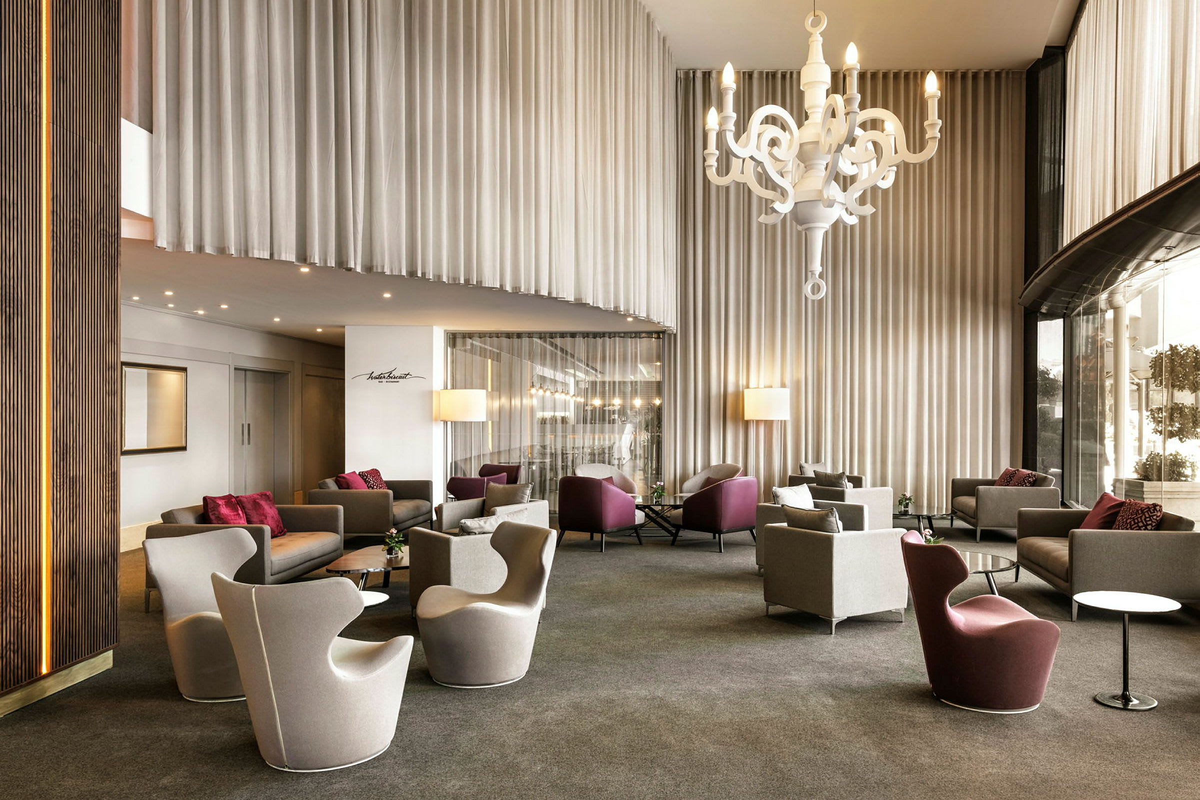 En hotellobby med moderna fåtöljer och soffor, ett stort vitt takhängande ljuskronelampställ och höga gardiner. (AI-genererad text)