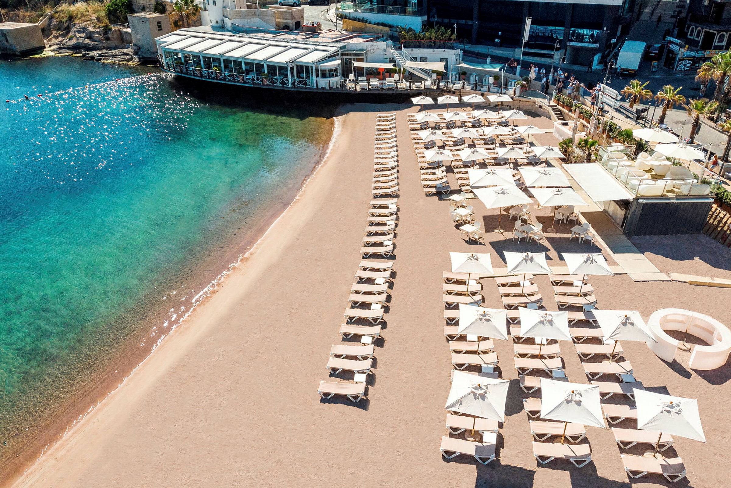 Strand med solstolar och parasoller samt en närliggande byggnad. (AI-genererad text)