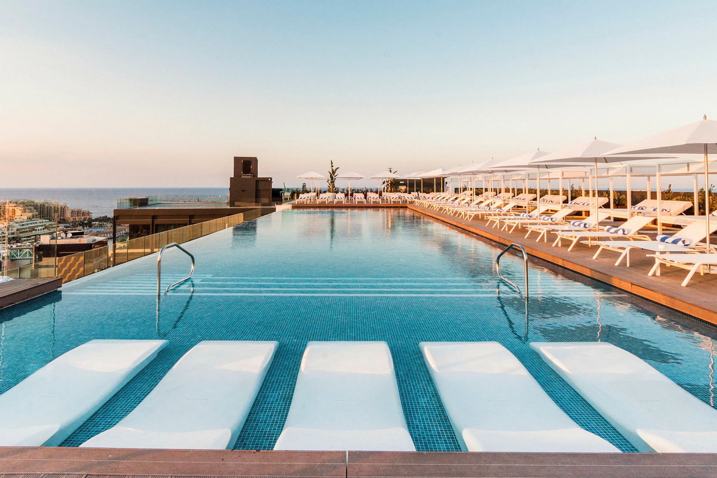 En infinitypool med solsängar och parasoller bredvid. Solnedgång syns i bakgrunden. (AI-genererad text)