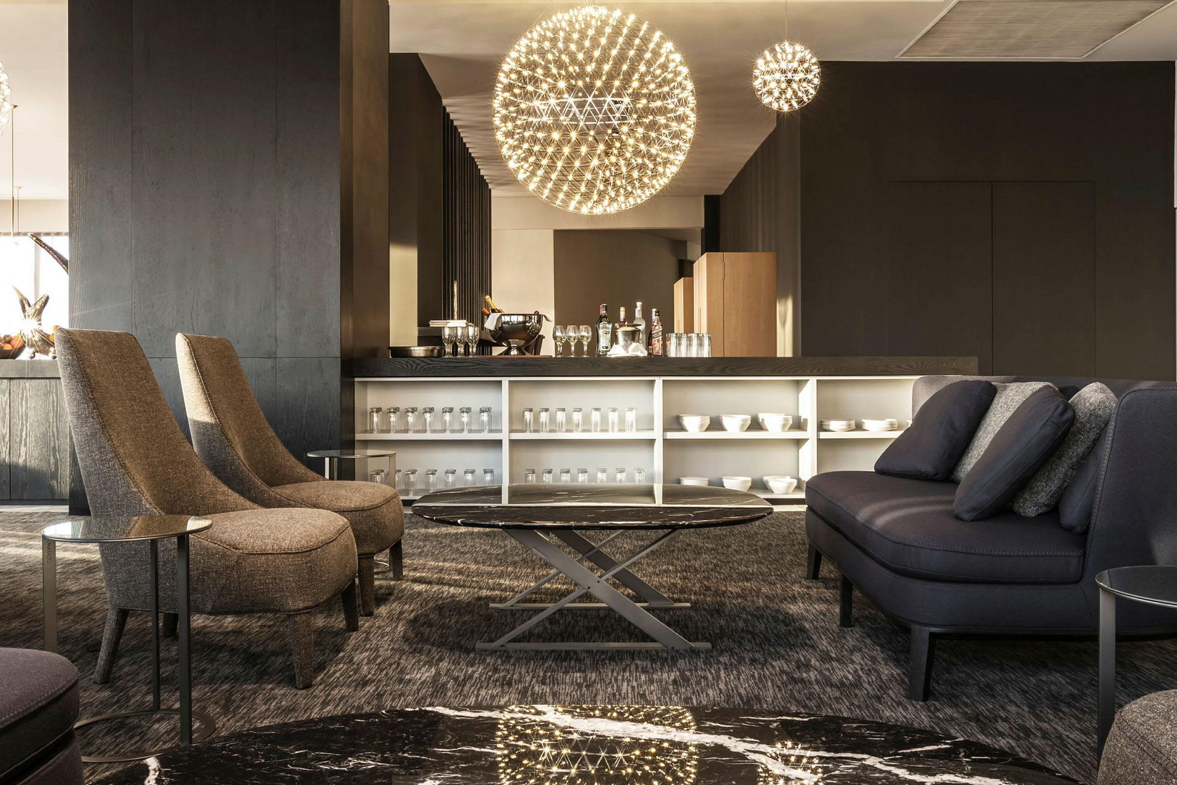 En modern lounge med kaffebar och sittgrupp med soffor och stolar. Utrymmet är välbelyst med unika hängande lampor och har ett elegant inrett bord. (AI-genererad text)
