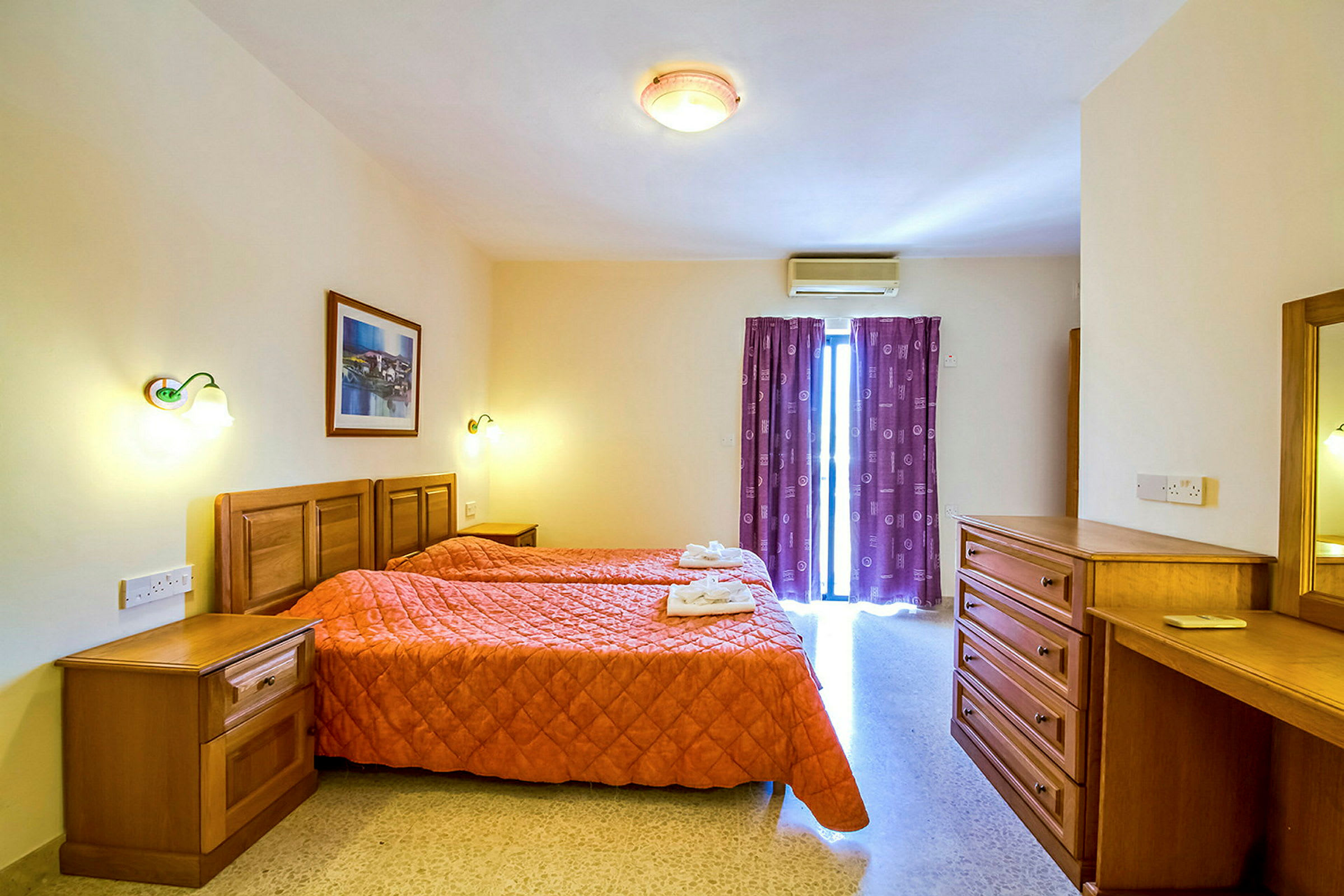 Ett hotellrum med två enkelsängar med orange täcken och mjuka mörklila gardiner framför ett fönster. Möbler av trä såsom en byrå, nattduksbord och skrivbord. (AI-genererad text)