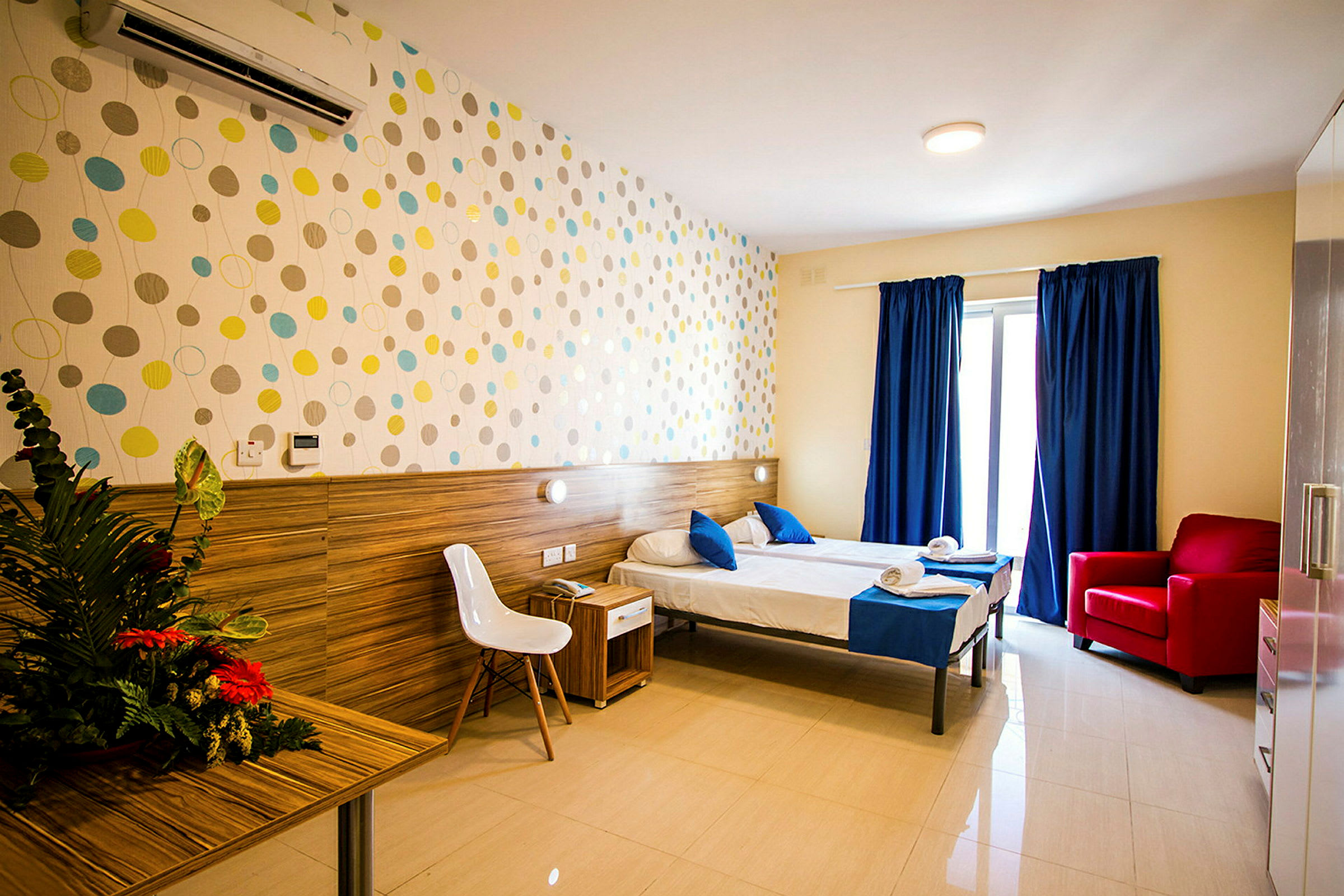Ett hotellrum med två enkelsängar, blå sängkläder, ett bord med blommor, en vit stol och en röd fåtölj. Färgglad dekoration pryder väggen. (AI-genererad text)