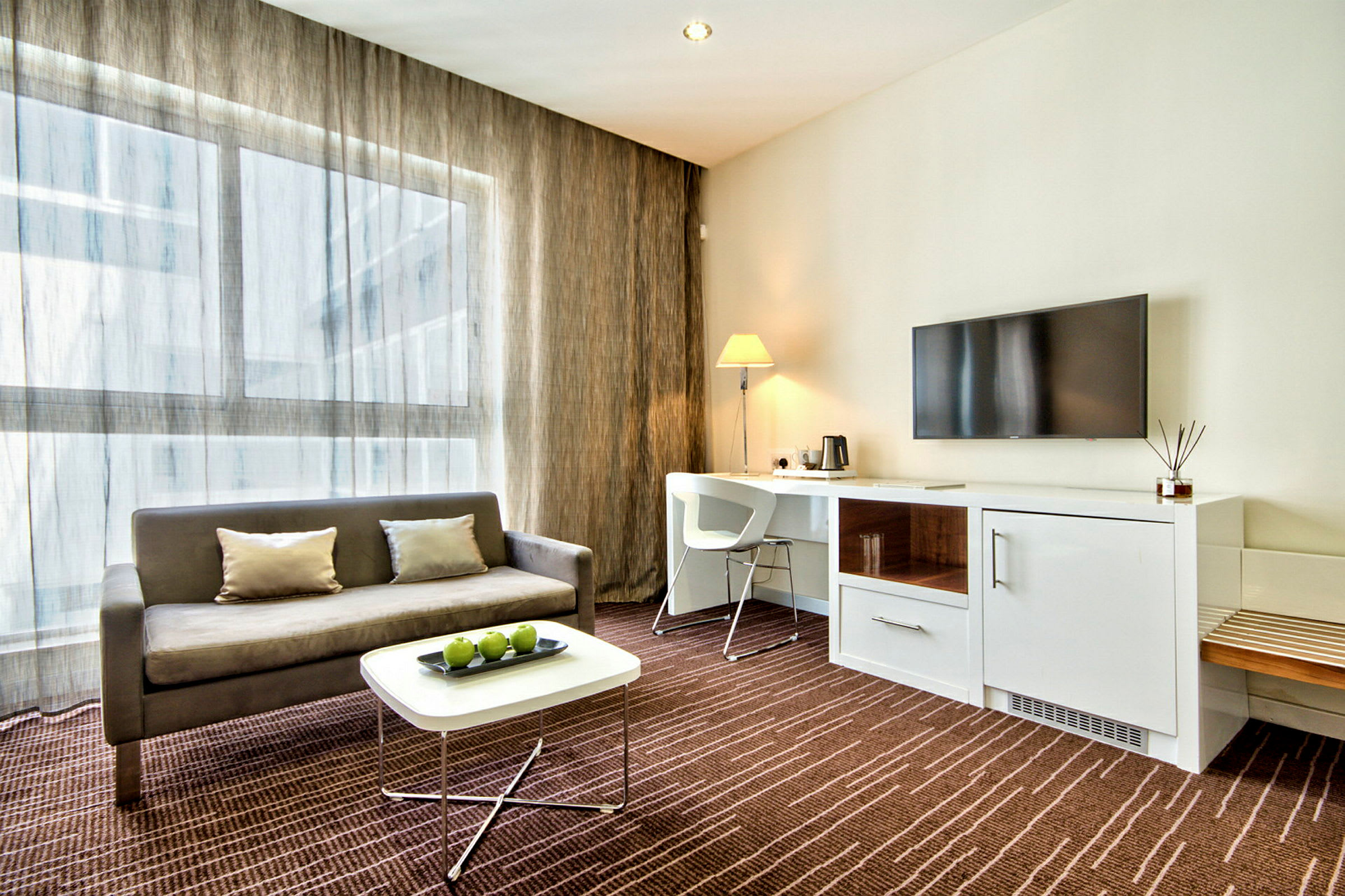 Ett modernt hotellrum med en soffa, ett soffbord med gröna äpplen, en arbetsyta med stol och lampa, samt en väggmonterad TV. (AI-genererad text)