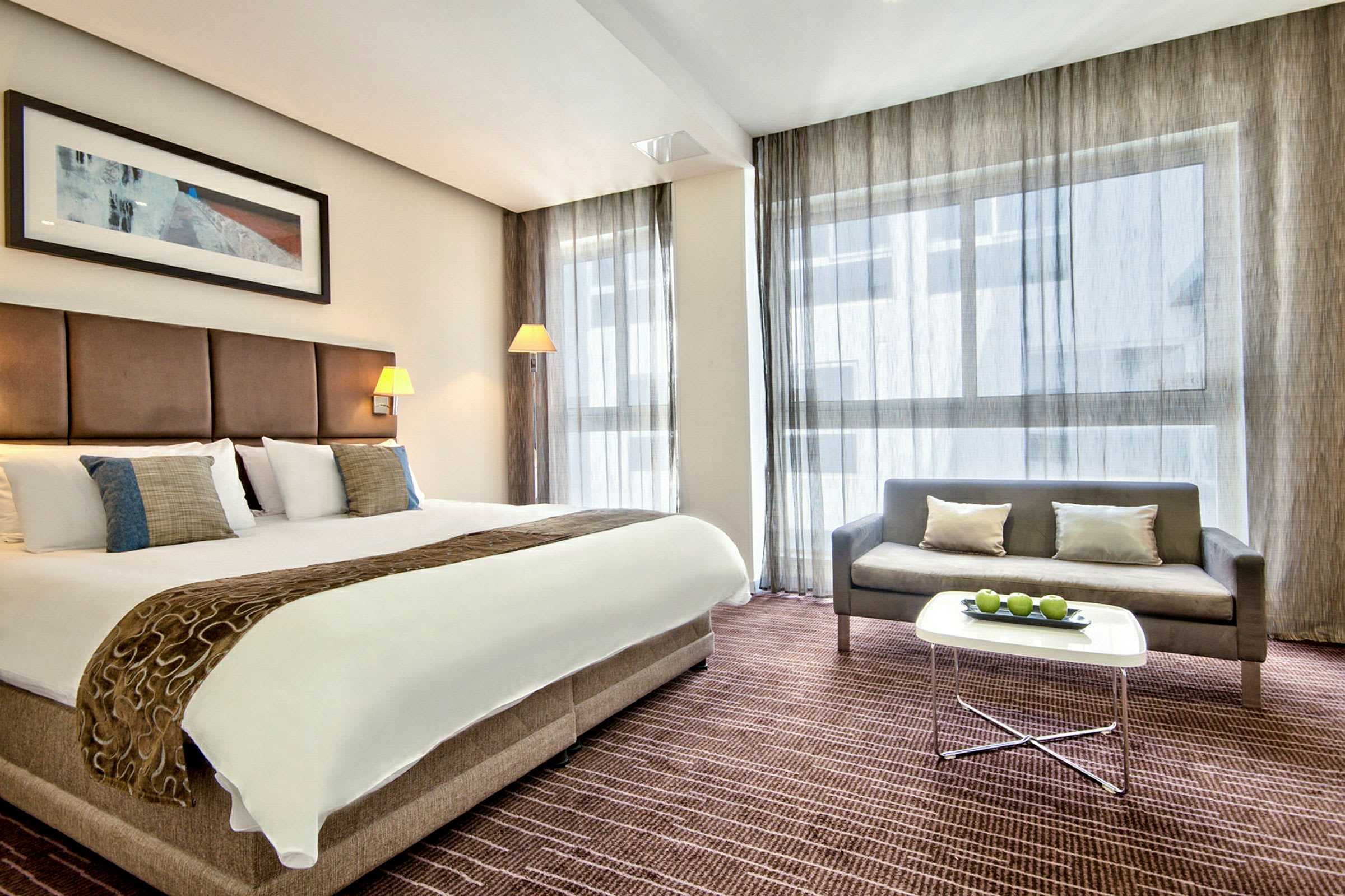 Ett hotellrum med en stor dubbelsäng, soffbord innehållande gröna äpplen och en grå soffa. (AI-genererad text)