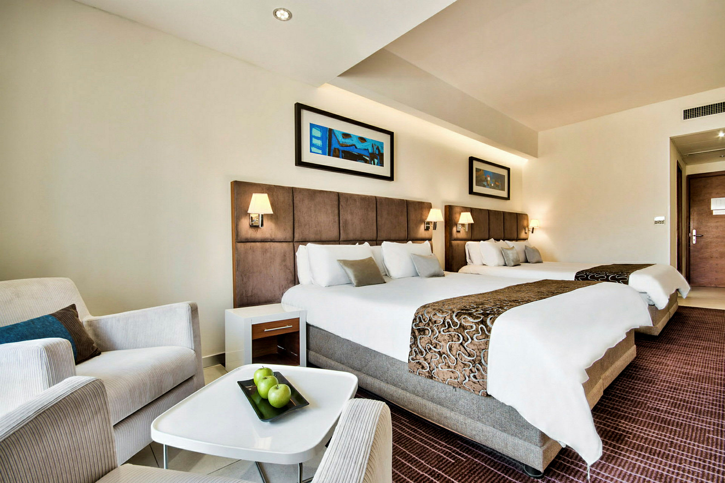 Ett hotellrum med två stora sängar utrustade med sänglampor, en fåtölj, ett litet bord med gröna äpplen och ett dörröppning i bakgrunden. (AI-genererad text)