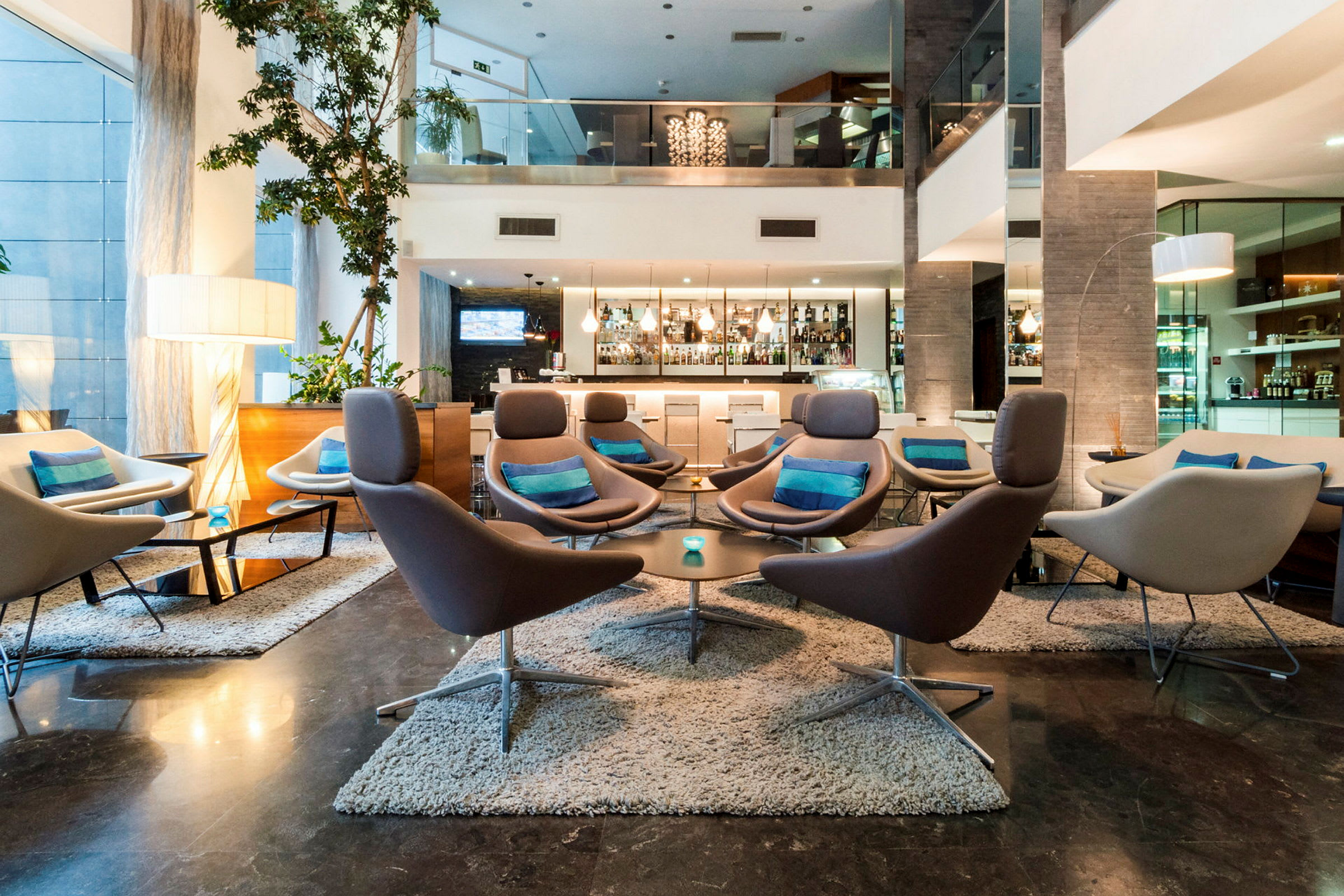 En hotellobby med moderna möbler, flera soffor och fåtöljer med kuddar i en stilren miljö. En bar syns i bakgrunden. (AI-genererad text)