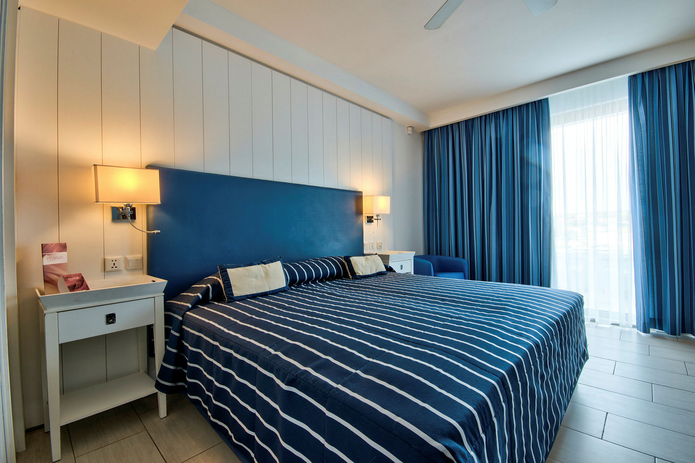 Ett hotellrum med en dubbelsäng klädd i blått och vitt randigt överkast, två sängbord och fönster med blå gardiner. (AI-genererad text)