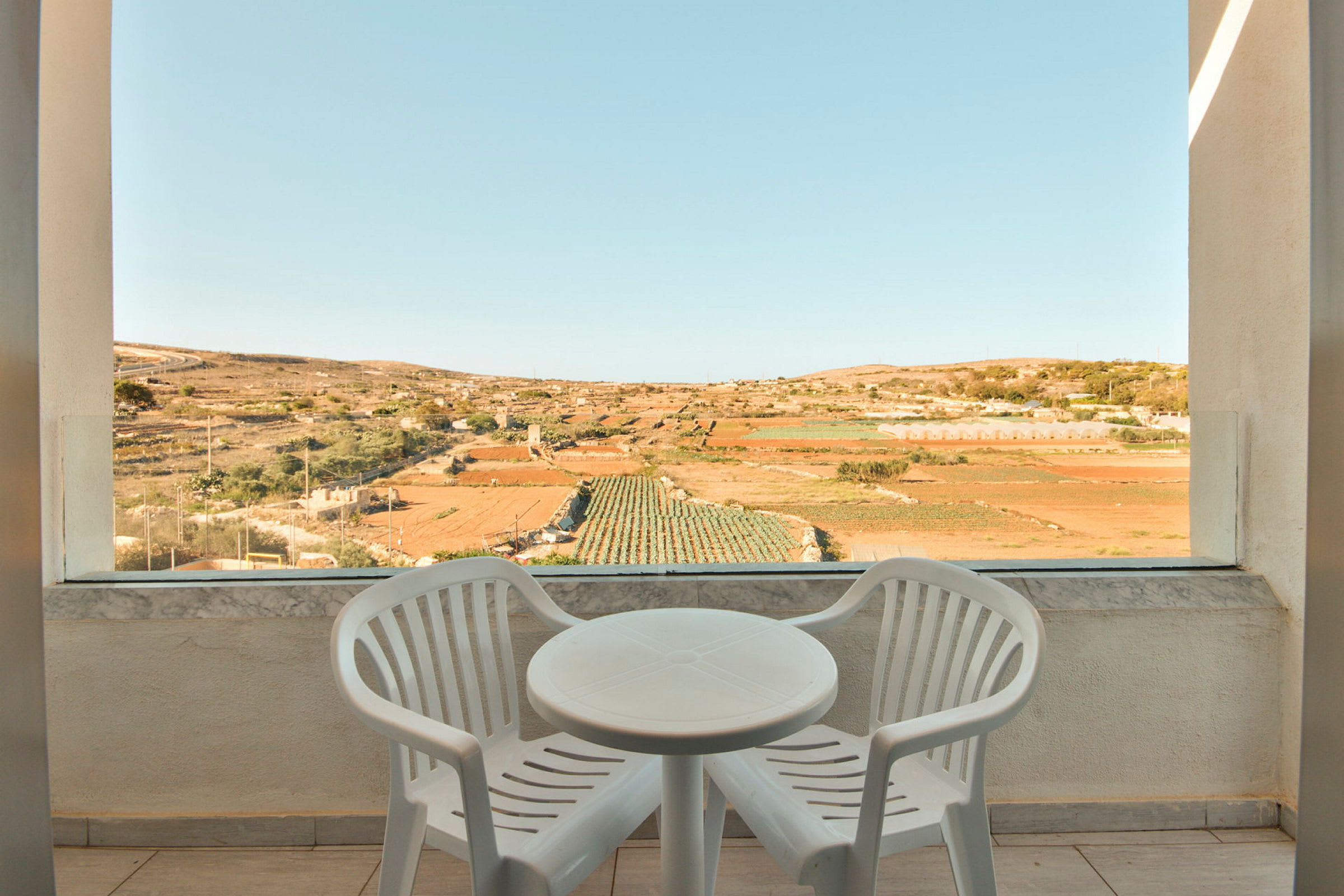 Två vita plaststolar och ett runt bord på en terrass med utsikt över odlingsfält. (AI-genererad text)