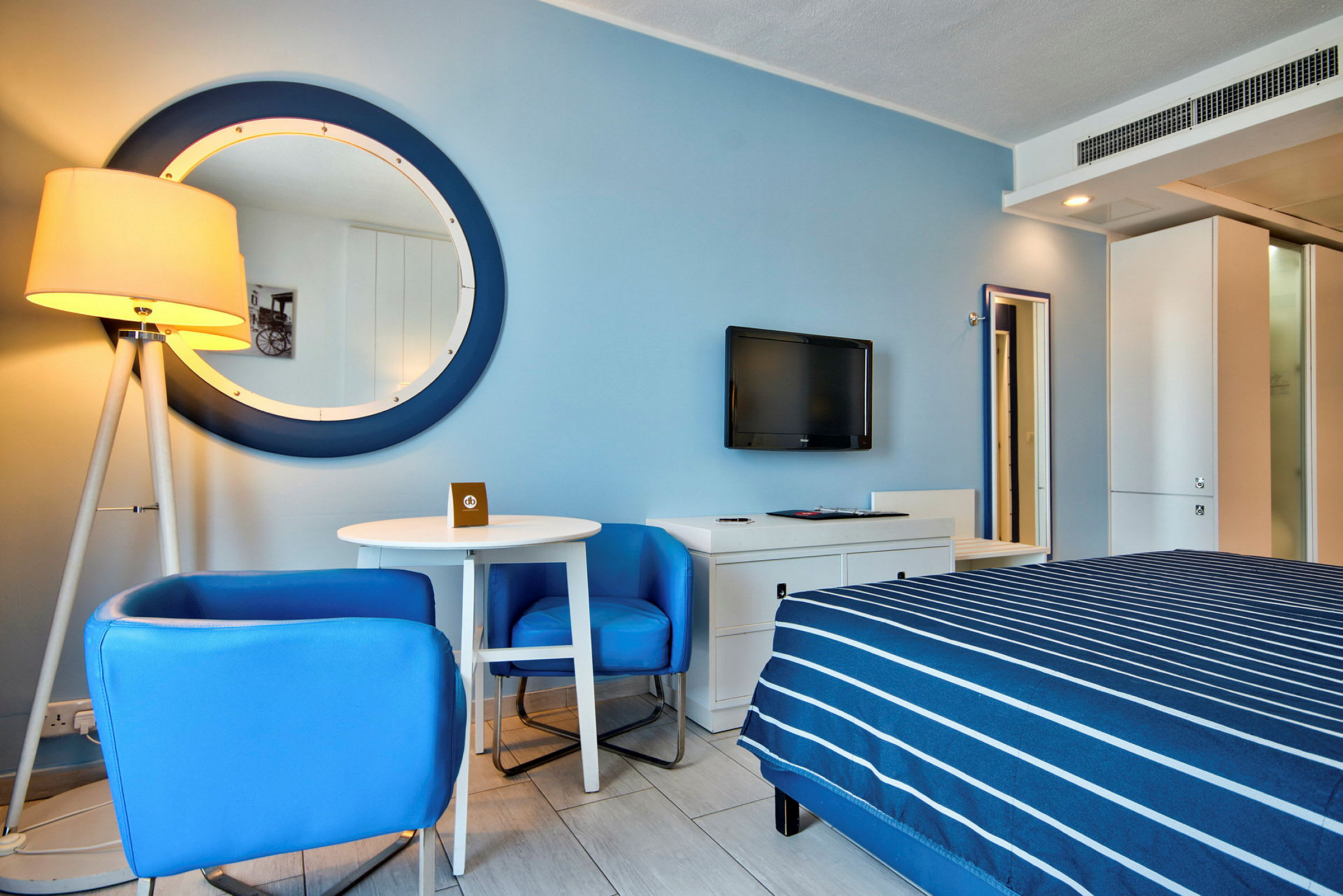 Ett hotellrum med blå väggar, en stor rund spegel, en säng med blå randigt överkast, ett vitt bord med blå stolar och en väggmonterad TV. (AI-genererad text)