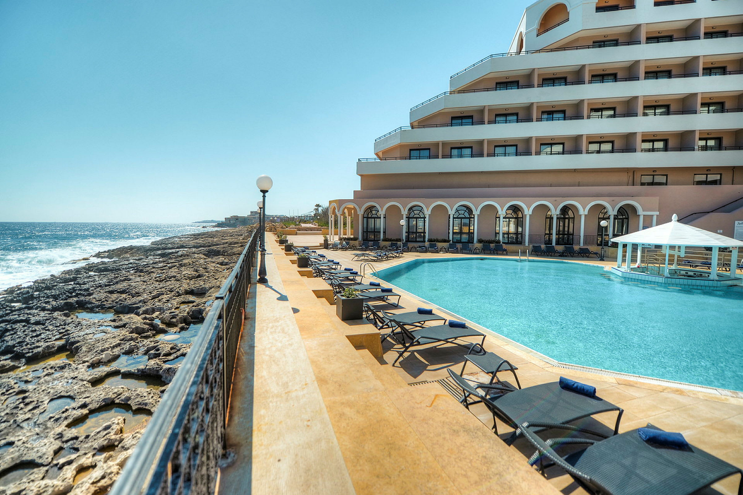 Bild som visar en lyxig resort vid havet med en swimmingpool, solstolar och en byggnad med balkonger. (AI-genererad text)