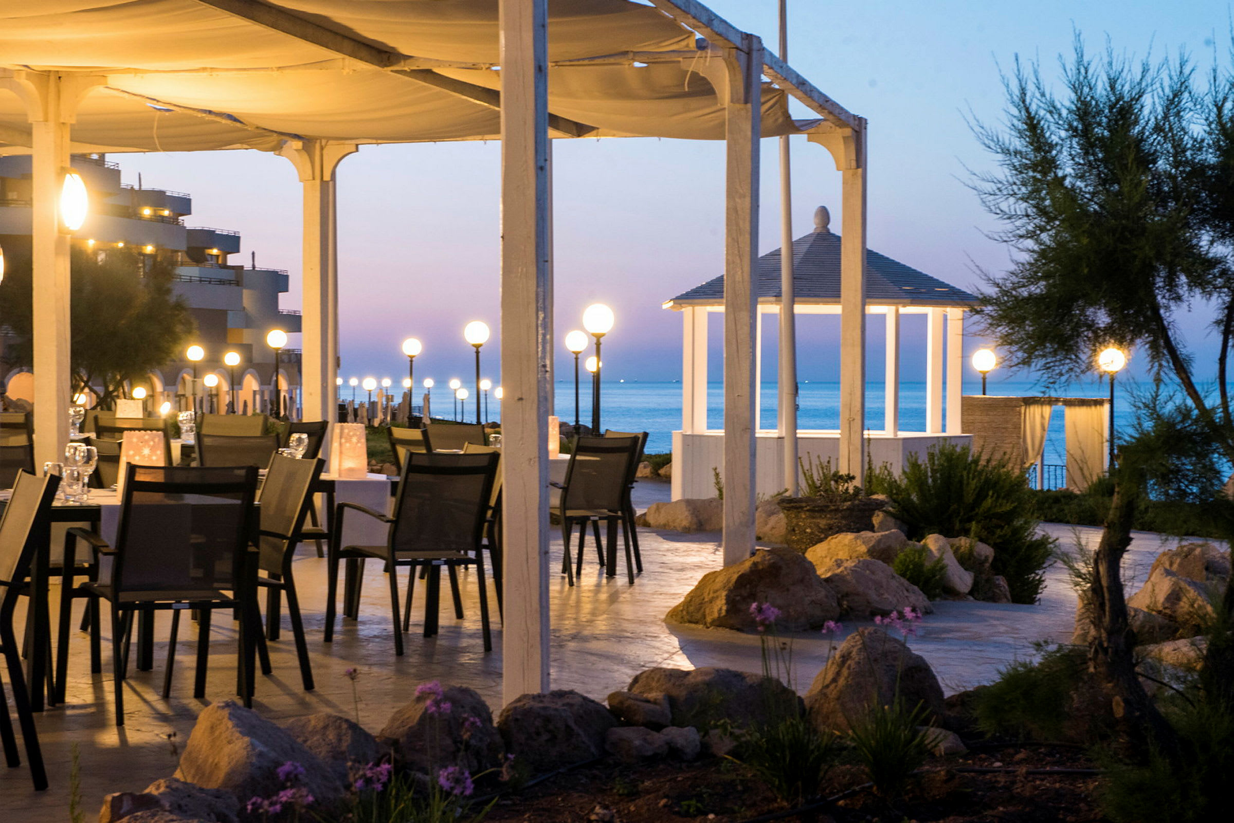 Ett upplyst utomhusområde med bord och stolar under ett tak nära havet. Lyktor skapar en behaglig atmosfär, och en vit paviljong syns mot bakgrunden. (AI-genererad text)